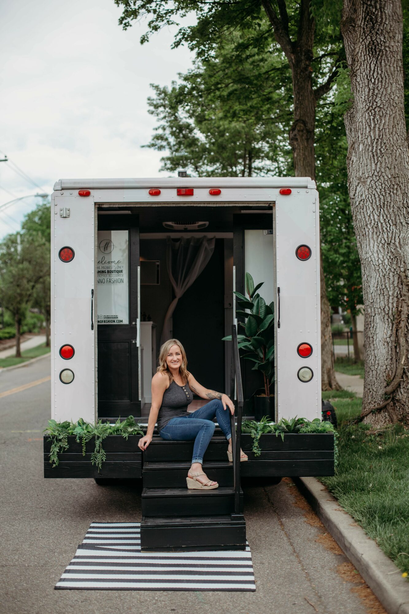 Woman sitting on steps in front of a mobile food truck, with trees and sidewalk nearby.