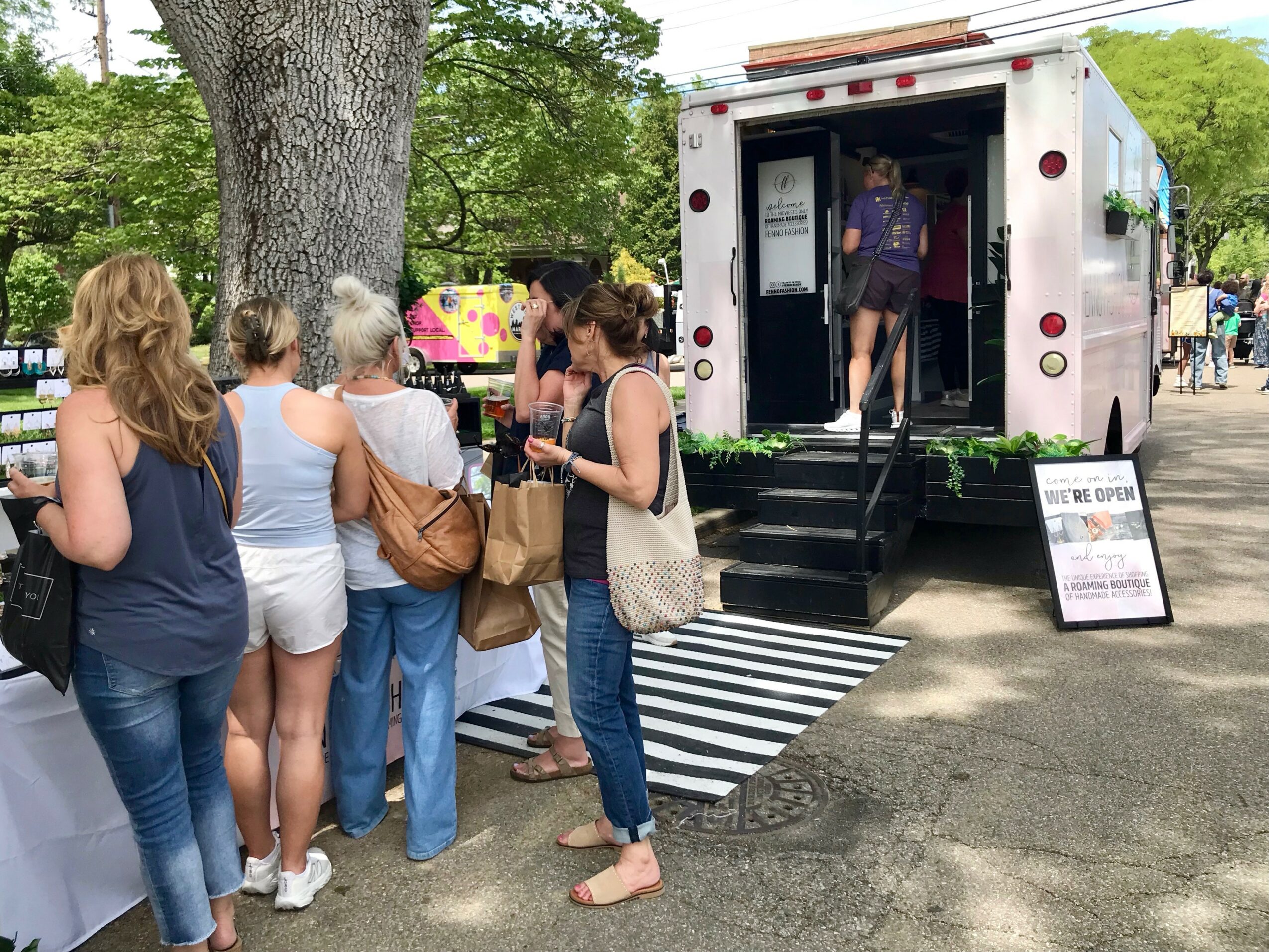 People gather outside a mobile food truck with a black and white striped mat, trees, and a signboard nearby.