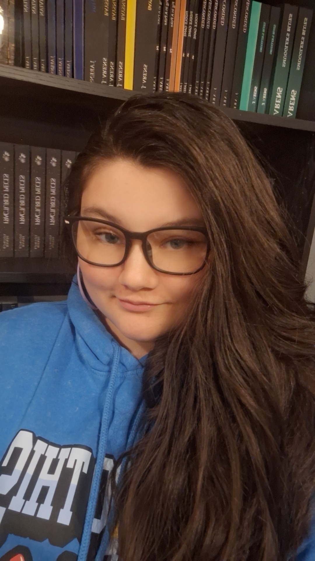 Young woman with glasses and long hair, wearing a blue hoodie, in front of a bookshelf.