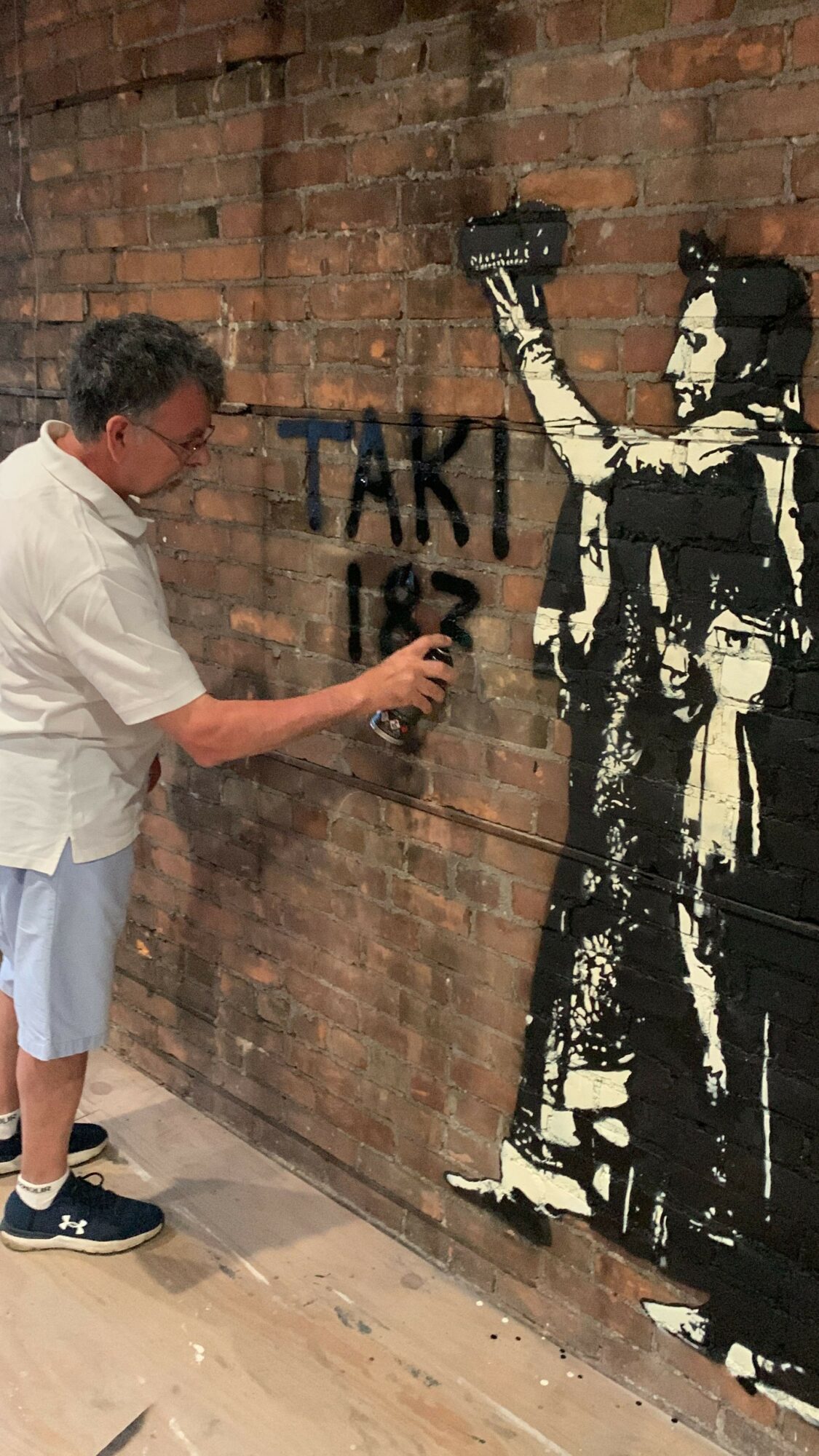 Person writing on a brick wall with graffiti, holding a spray can, in a room with wooden flooring.