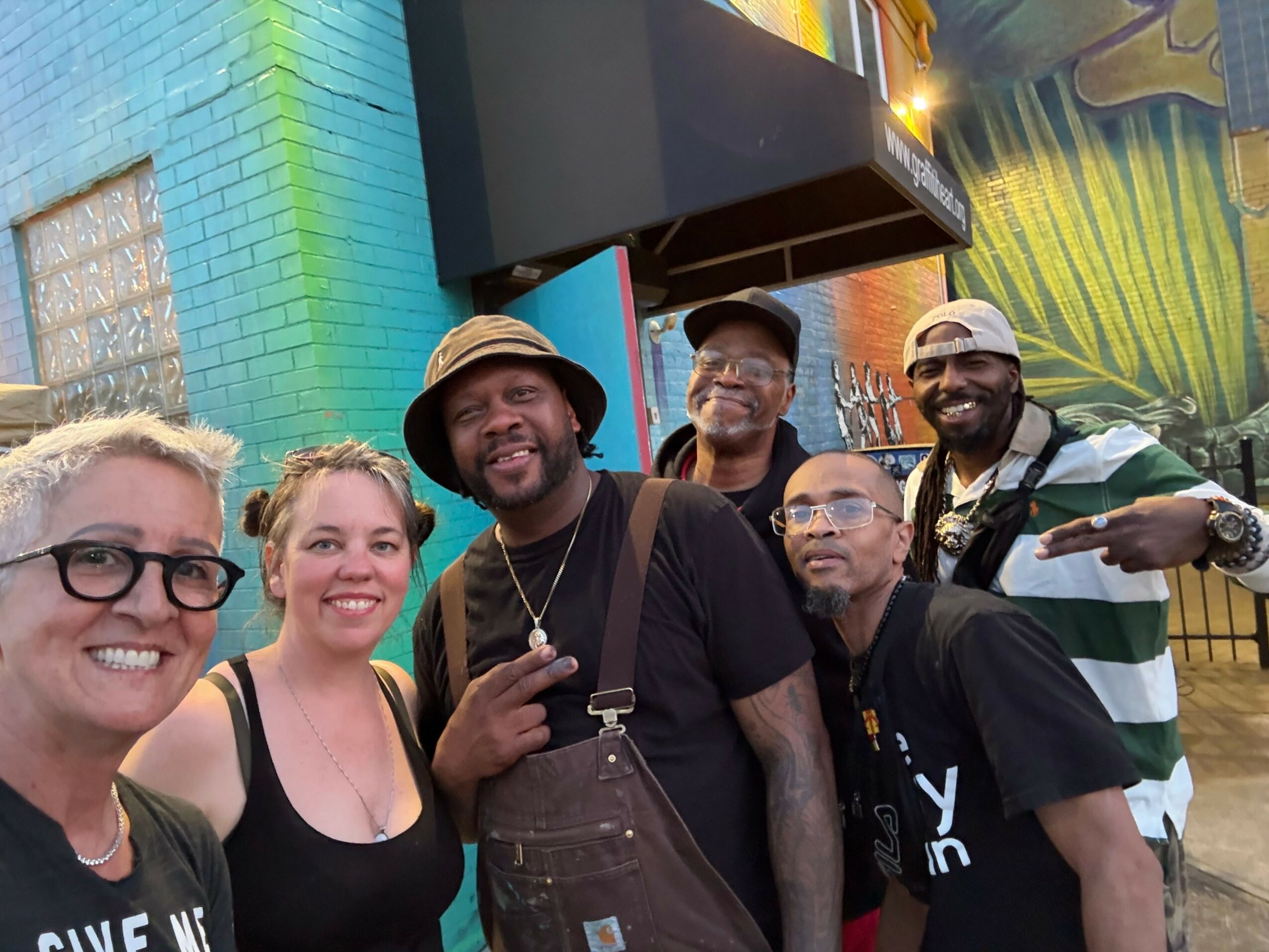 Group of seven people standing outdoors, smiling, with a colorful brick wall in the background.