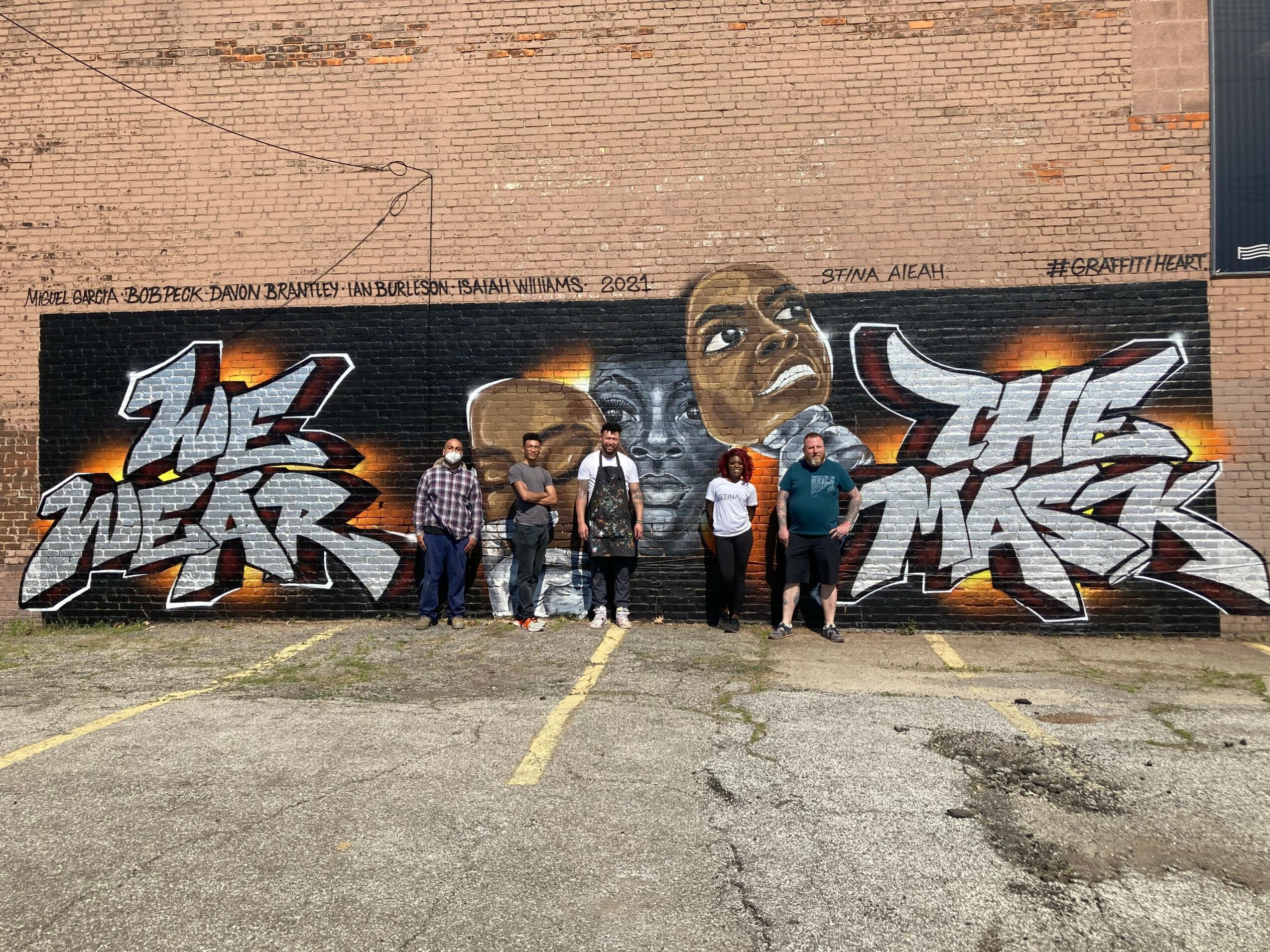 Five people stand in front of a graffiti mural with two large stylized text pieces and two faces, on a brick wall.