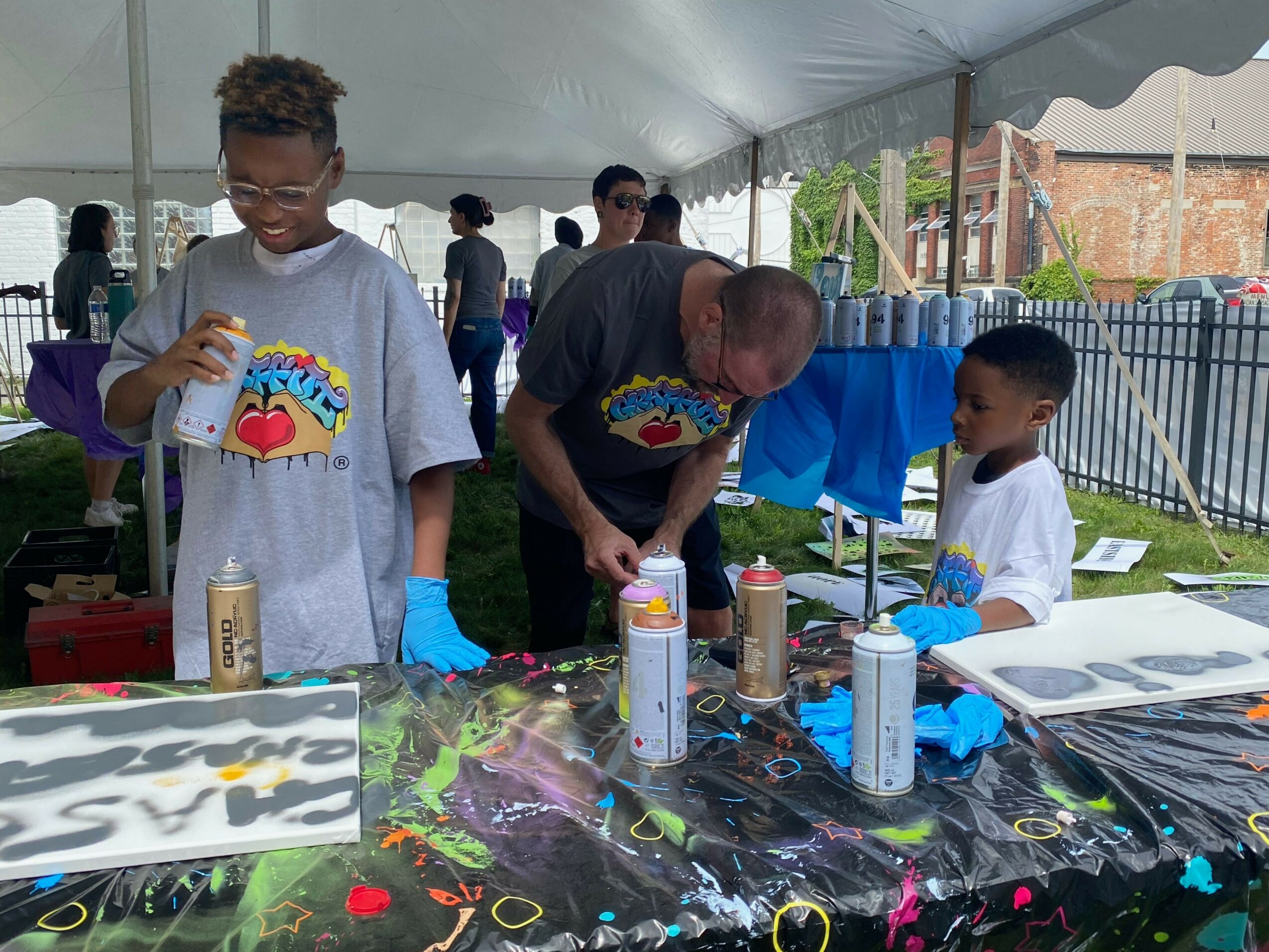 People at a table with spray paint cans, outdoors, with a tent and fence in background.