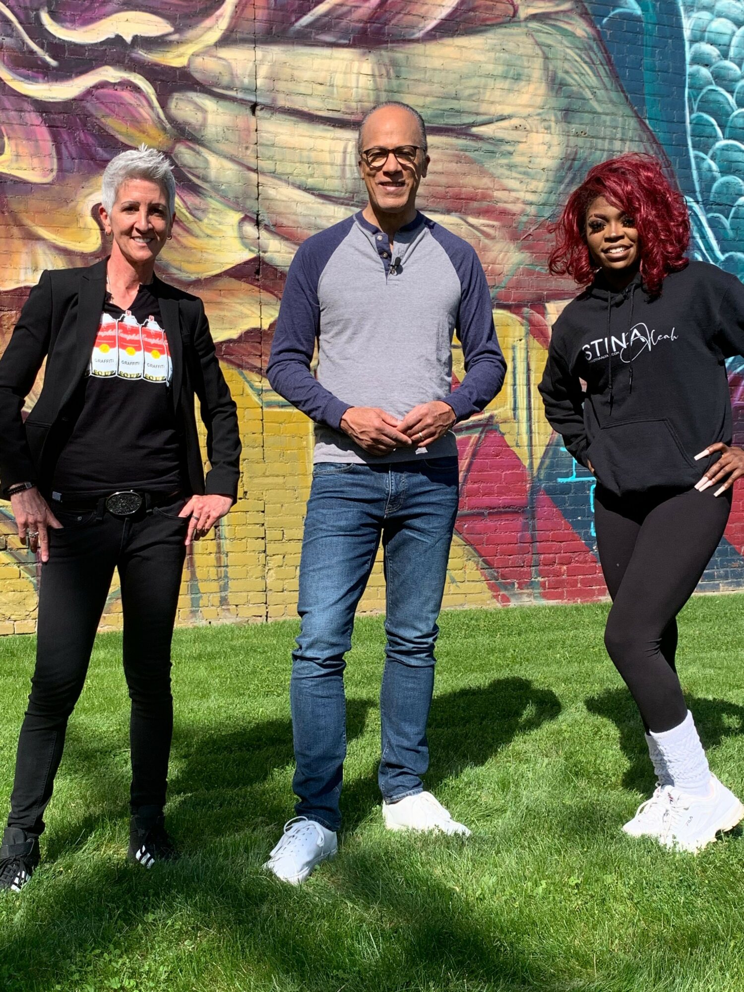 Three people standing outdoors on grass in front of a colorful mural, posing for a photo.