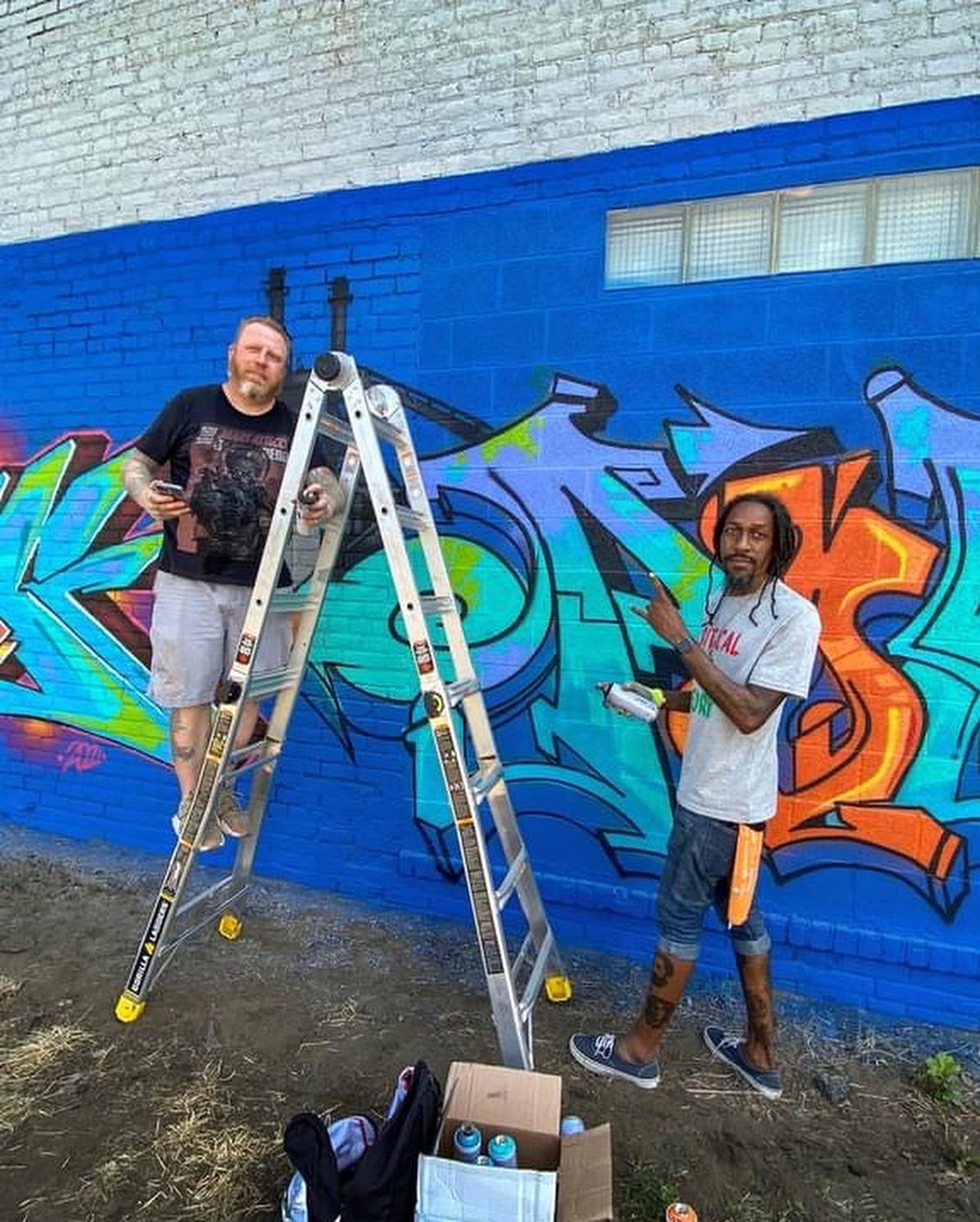 Two men stand in front of a colorful graffiti wall, one on a ladder and the other holding a spray can, outdoors.