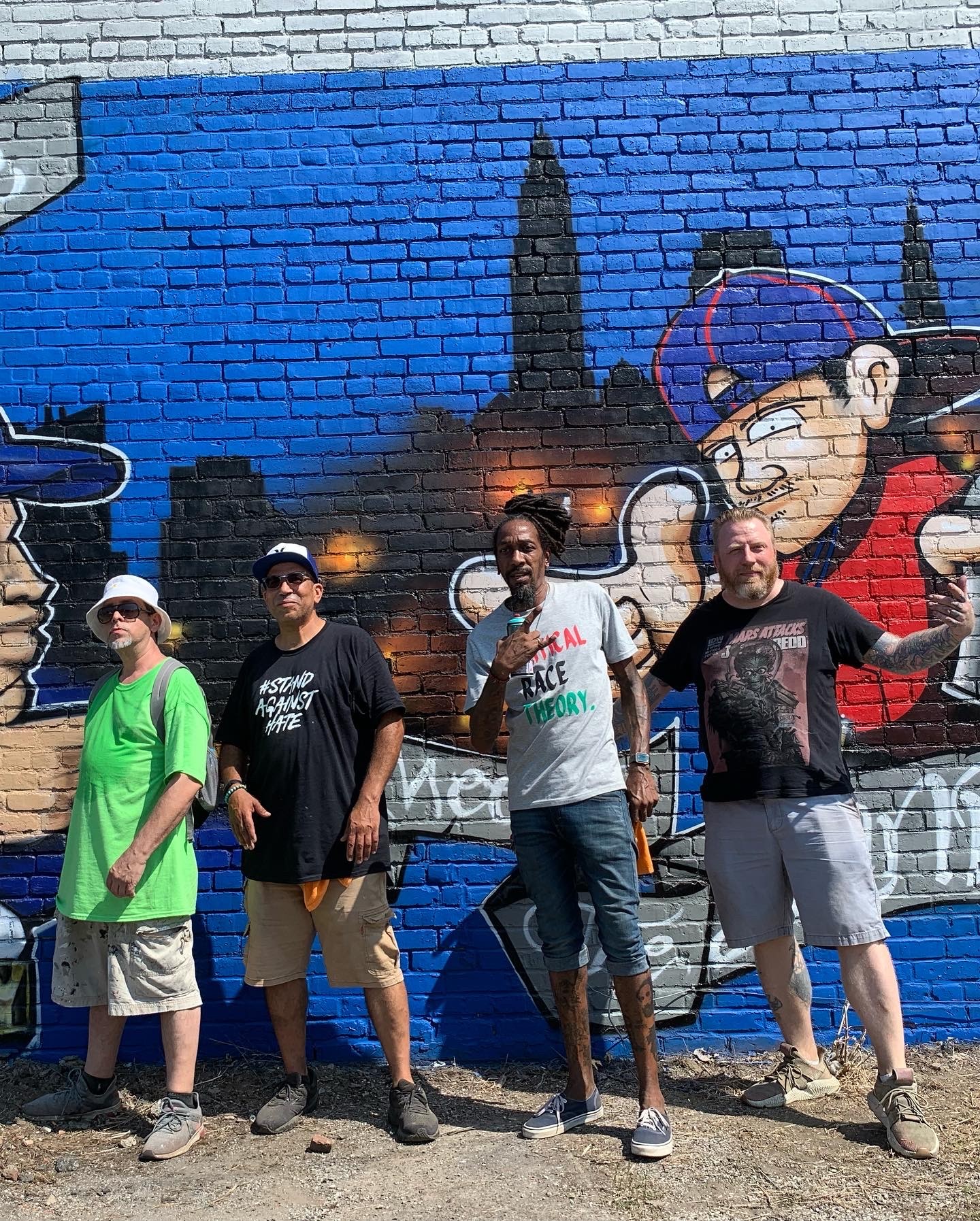 Four people stand in front of a colorful mural of cartoon characters and city skyline, holding a banner.