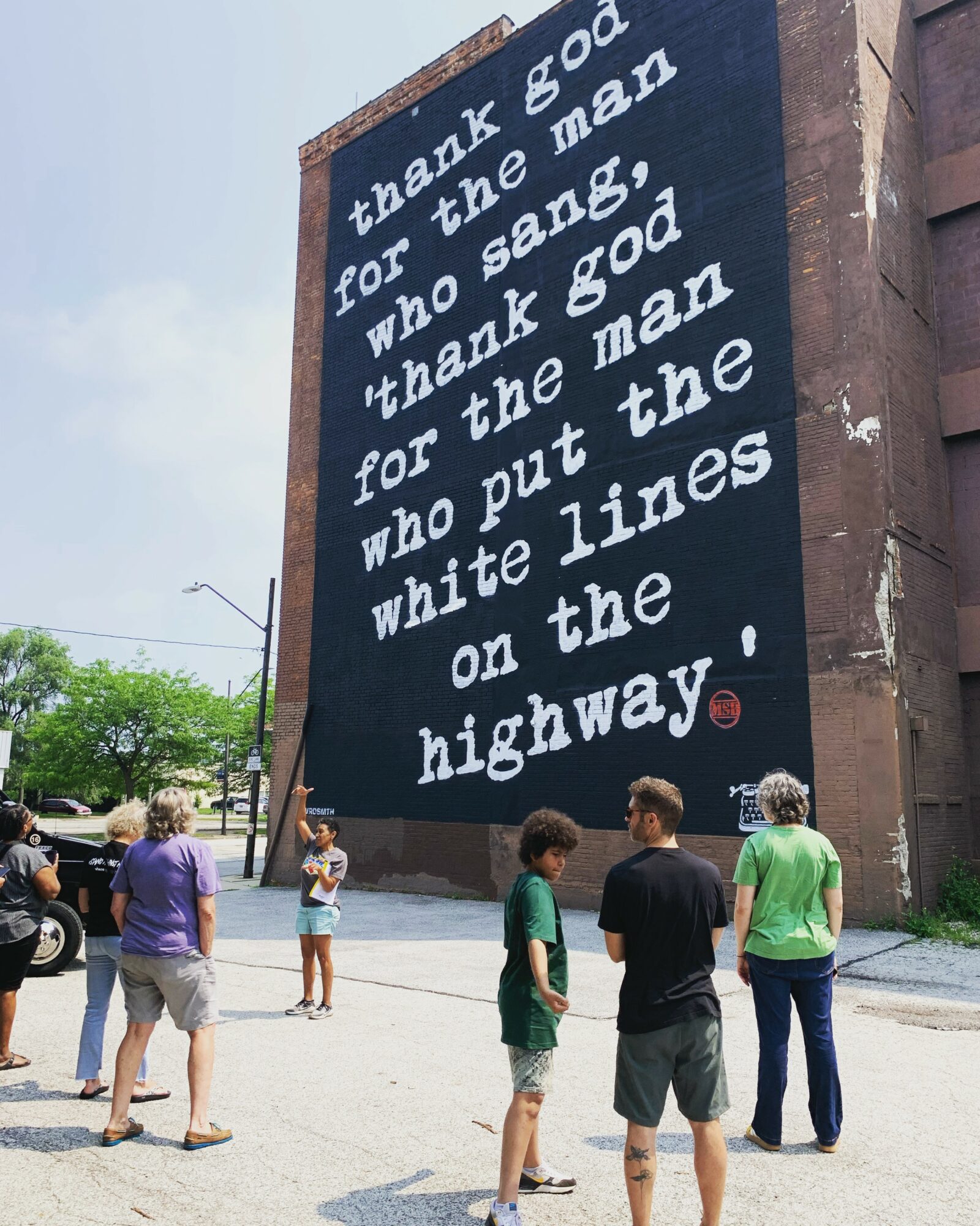 Large mural with white text on a dark background, with people standing and walking nearby outdoors.