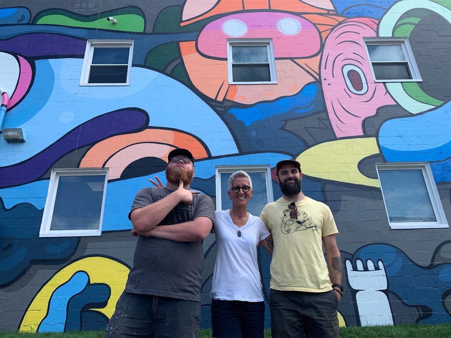 Three people stand in front of a colorful, abstract mural with windows on a building facade.