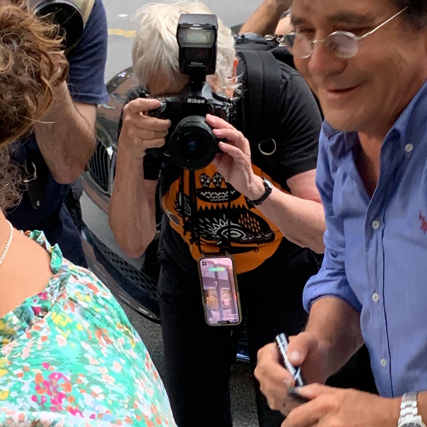 People gathered outdoors, one person taking a photo with a camera, others engaged in conversation, some holding pens, colorful clothing visible.