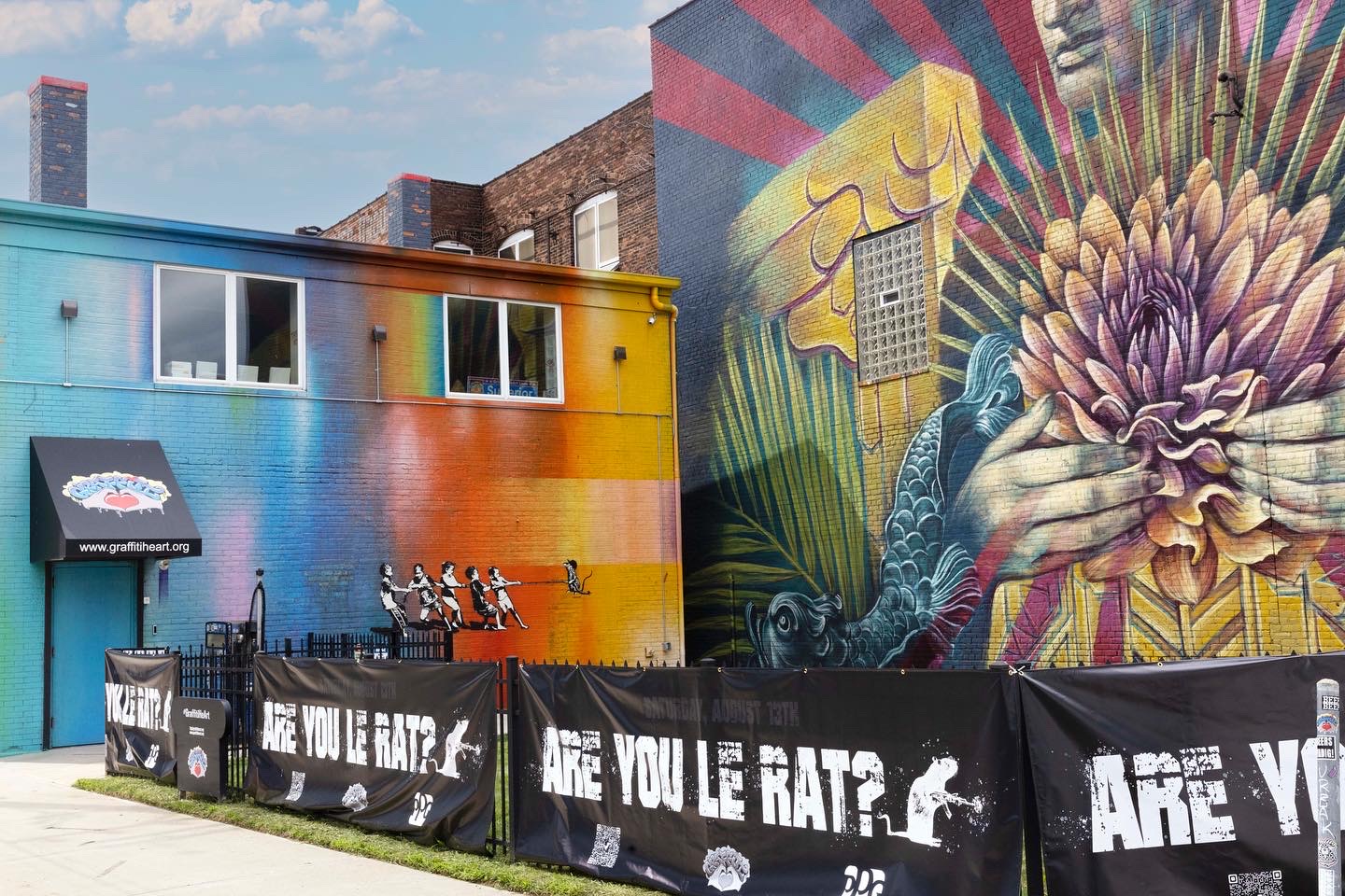 Colorful mural on building wall with abstract and floral designs, black fencing with white text, and a small awning on the left.