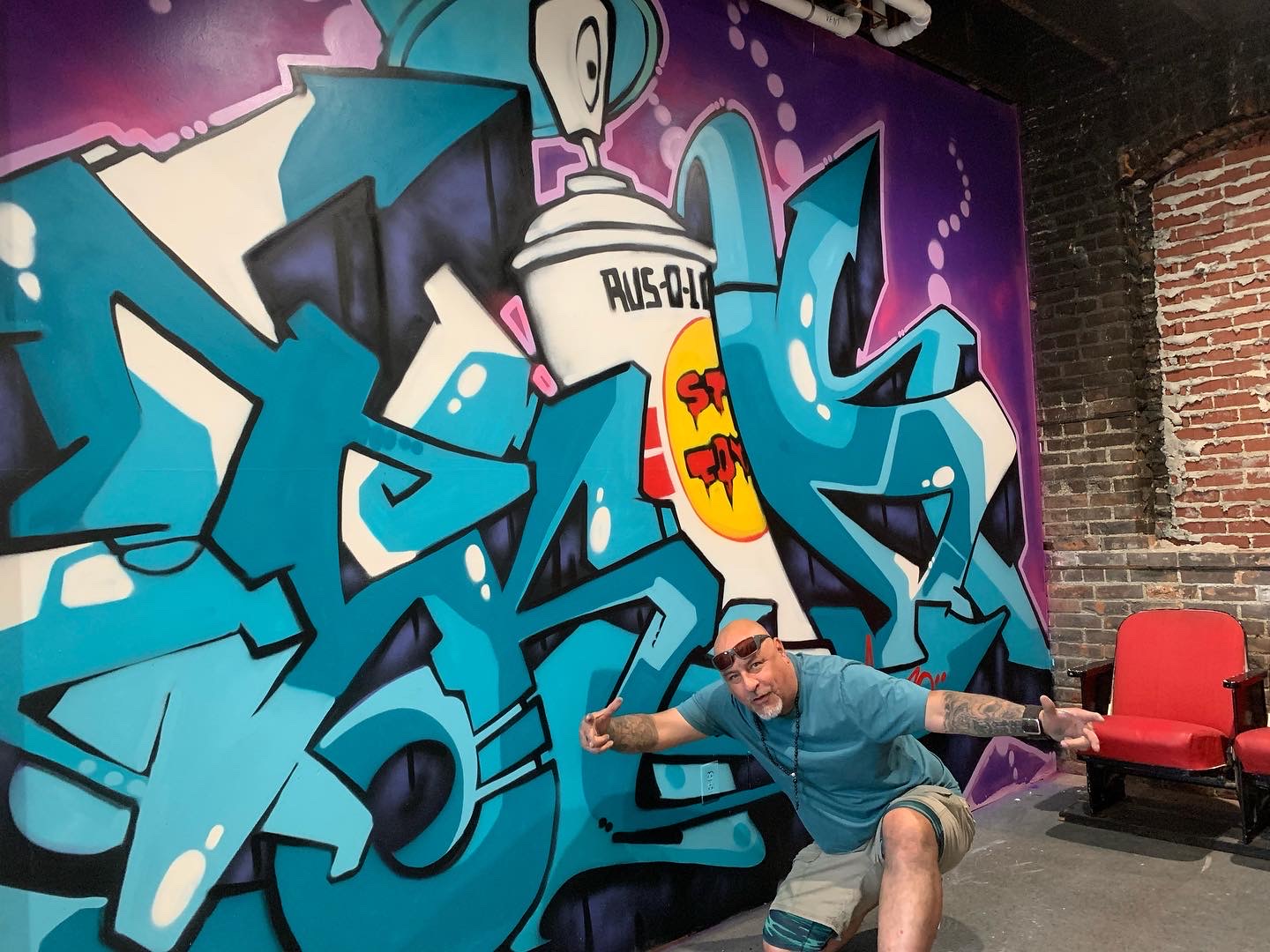 Person in blue shirt and shorts sitting in front of colorful graffiti wall, with red chairs nearby.