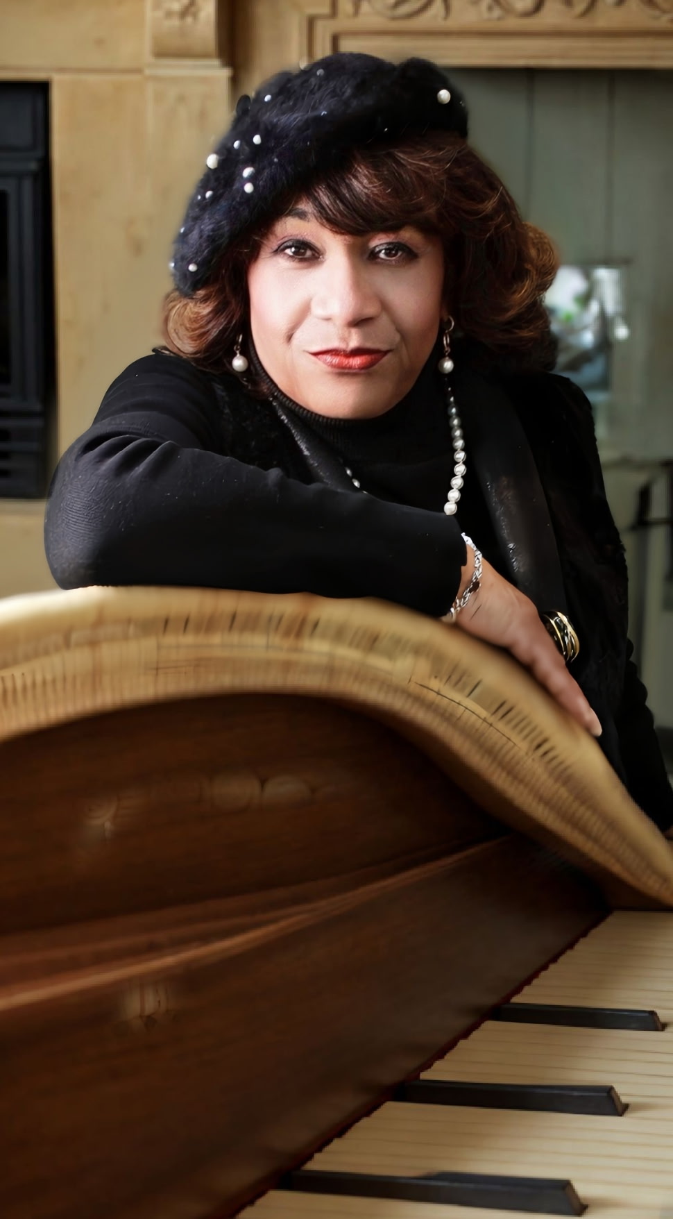 Woman with dark hair, wearing a black hat and pearl necklace, sitting at a piano with a wooden interior background.