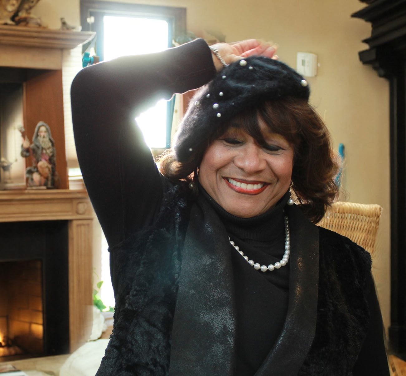 Woman smiling, wearing a black outfit and pearl necklace, with her arm raised, indoors with a fireplace and decorations.