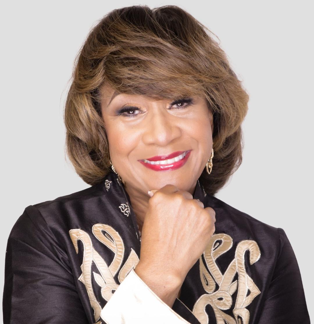 Smiling woman with short hair, wearing a black jacket with gold embroidery, resting her chin on her hand.