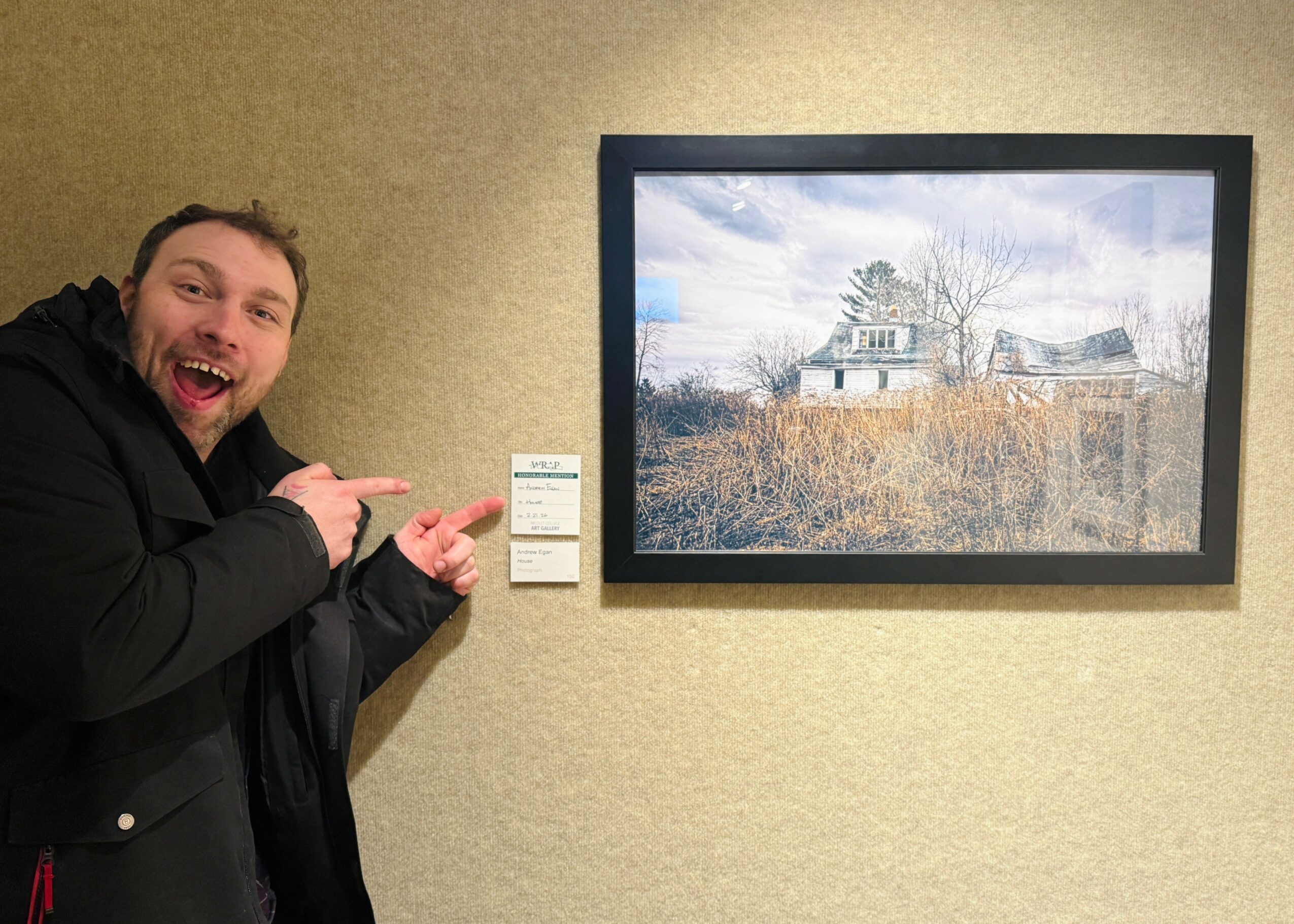 Man smiling and pointing at a framed landscape photograph on a beige wall.