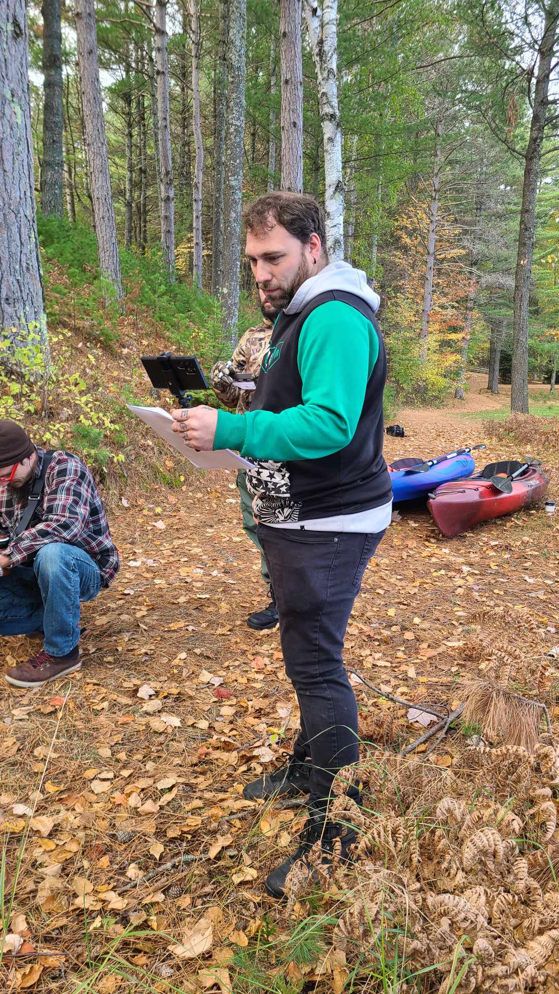 Man standing outdoors in a forest, reading a map or paper, with trees and fallen leaves around him.