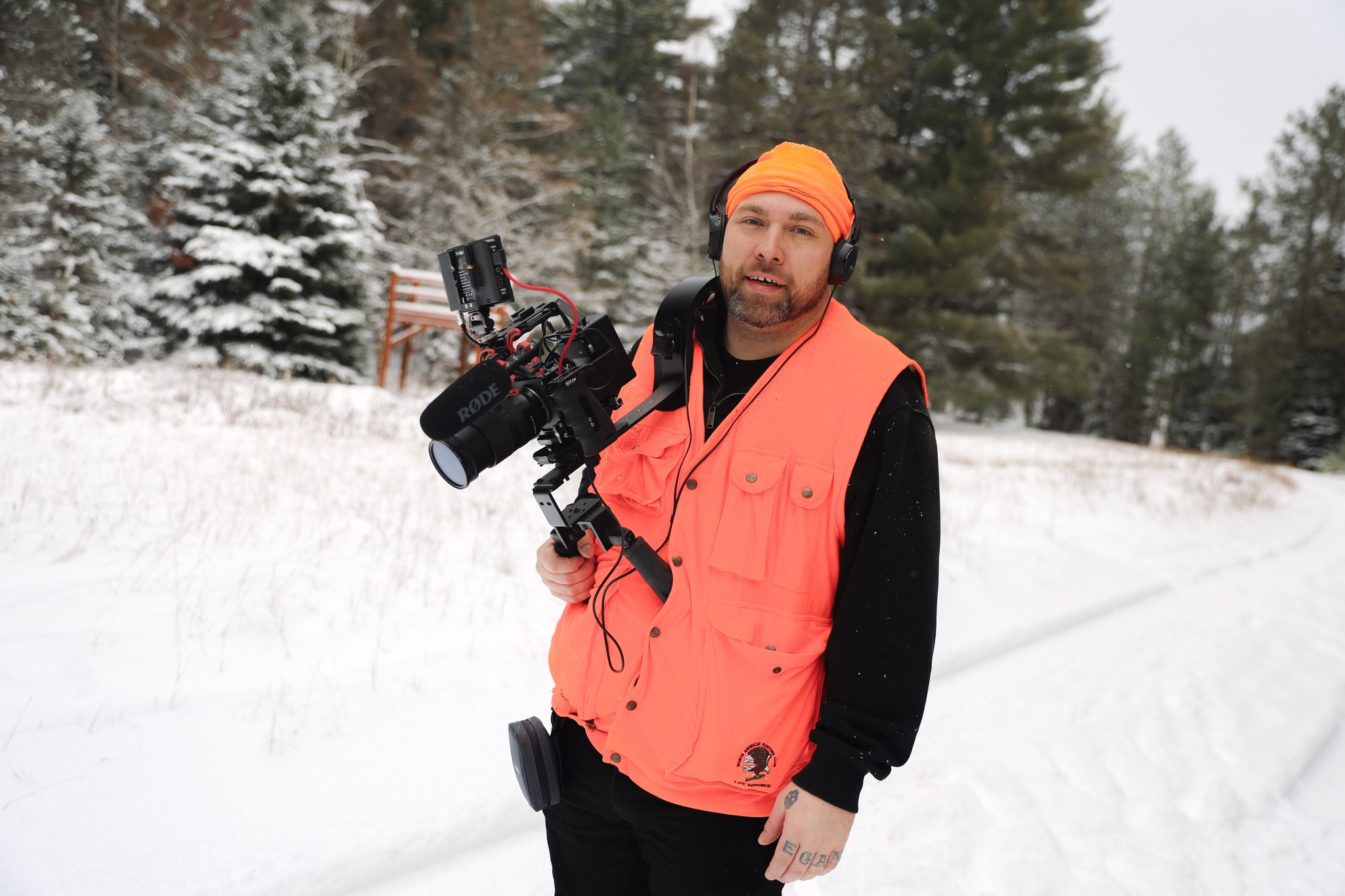 Man in orange vest and beanie holding a camera in snowy outdoor setting with trees