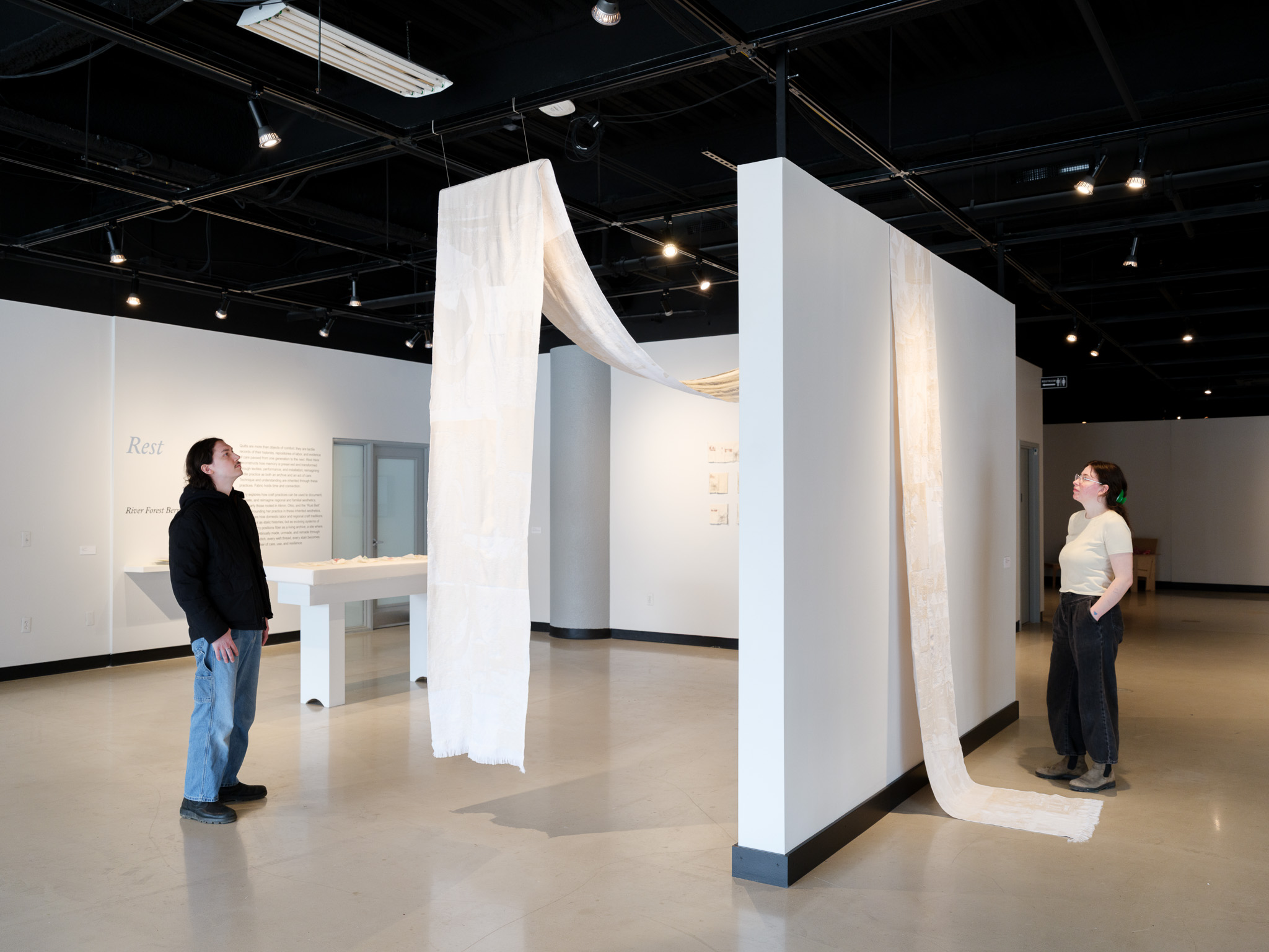 Two people stand in an art gallery near a white installation with hanging fabric and paper-like elements, in a spacious room.