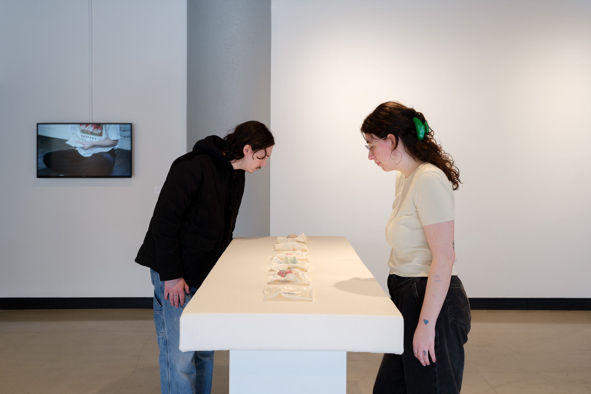 Two women stand at a white table examining artwork in a gallery, with a wall-mounted screen in the background.
