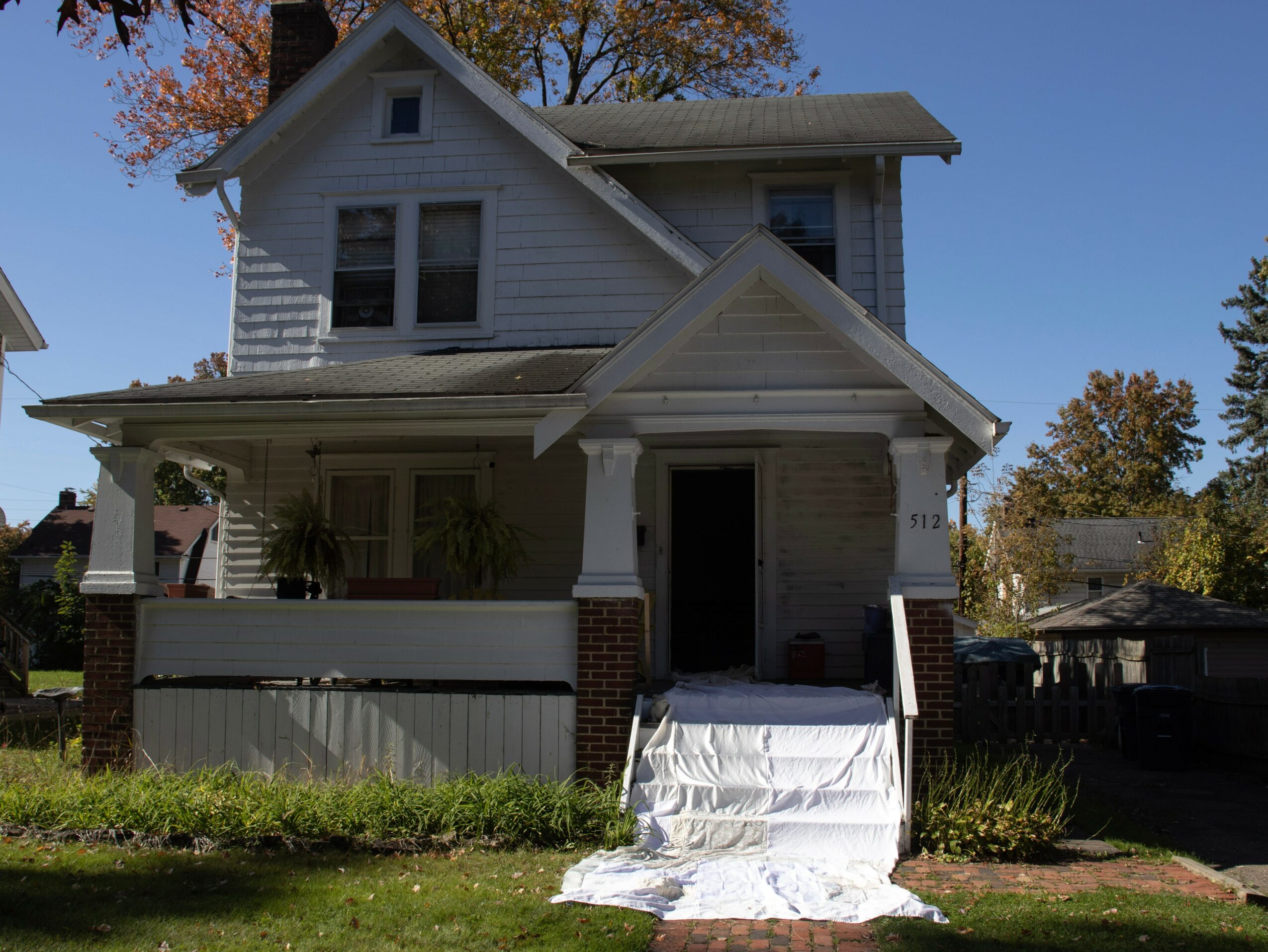 Two-story house with front porch, stairs covered with white cloth, and a small yard, under a clear blue sky.