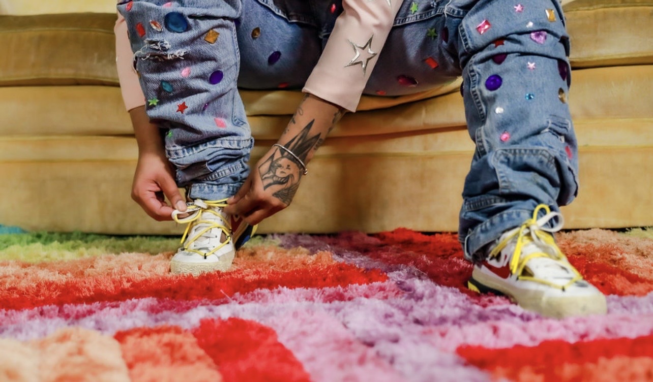 Person tying shoelaces on a colorful rug, wearing blue jeans with colorful dots, yellow shoes, and a tattooed arm, sitting on a yellow couch.