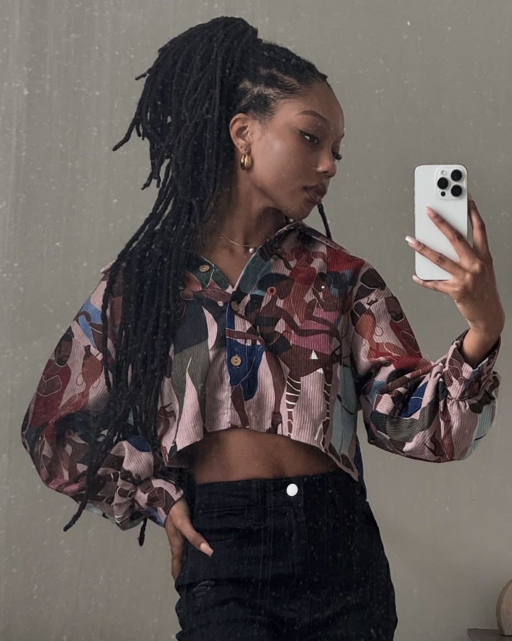 Young woman with dreadlocks taking a mirror selfie, wearing a cropped patterned jacket and high-waisted pants.