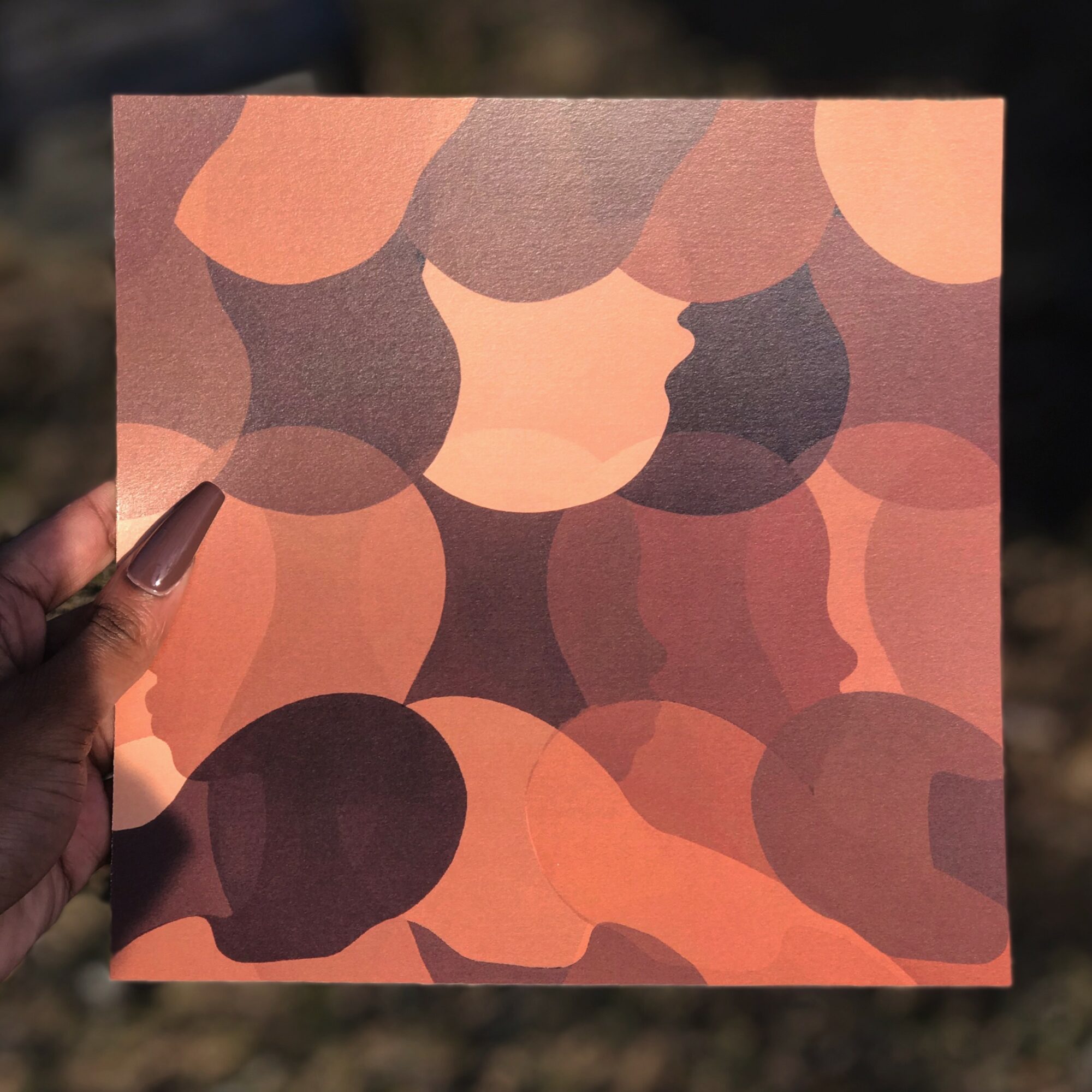 A person holds a square piece of paper with an abstract pattern of overlapping shapes in shades of pink, purple, and peach.