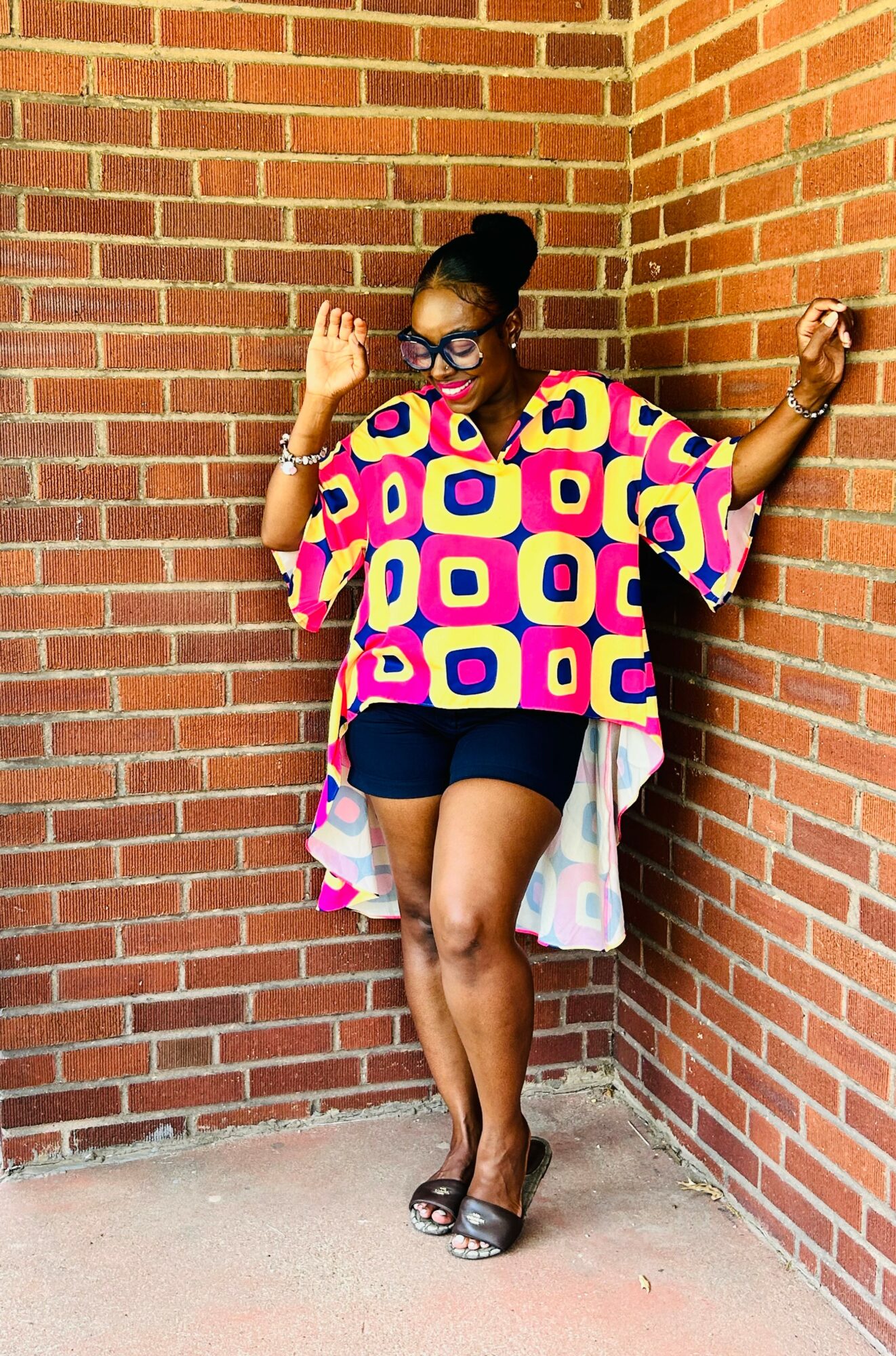 Woman in colorful geometric top, black shorts, glasses, and sandals leaning against brick wall outdoors.