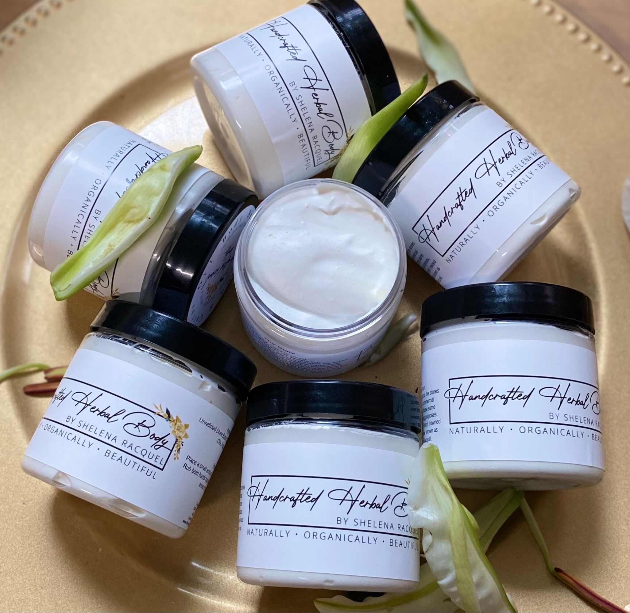 Seven jars of cream arranged on a gold tray with green leaves, some jars open showing white cream inside.
