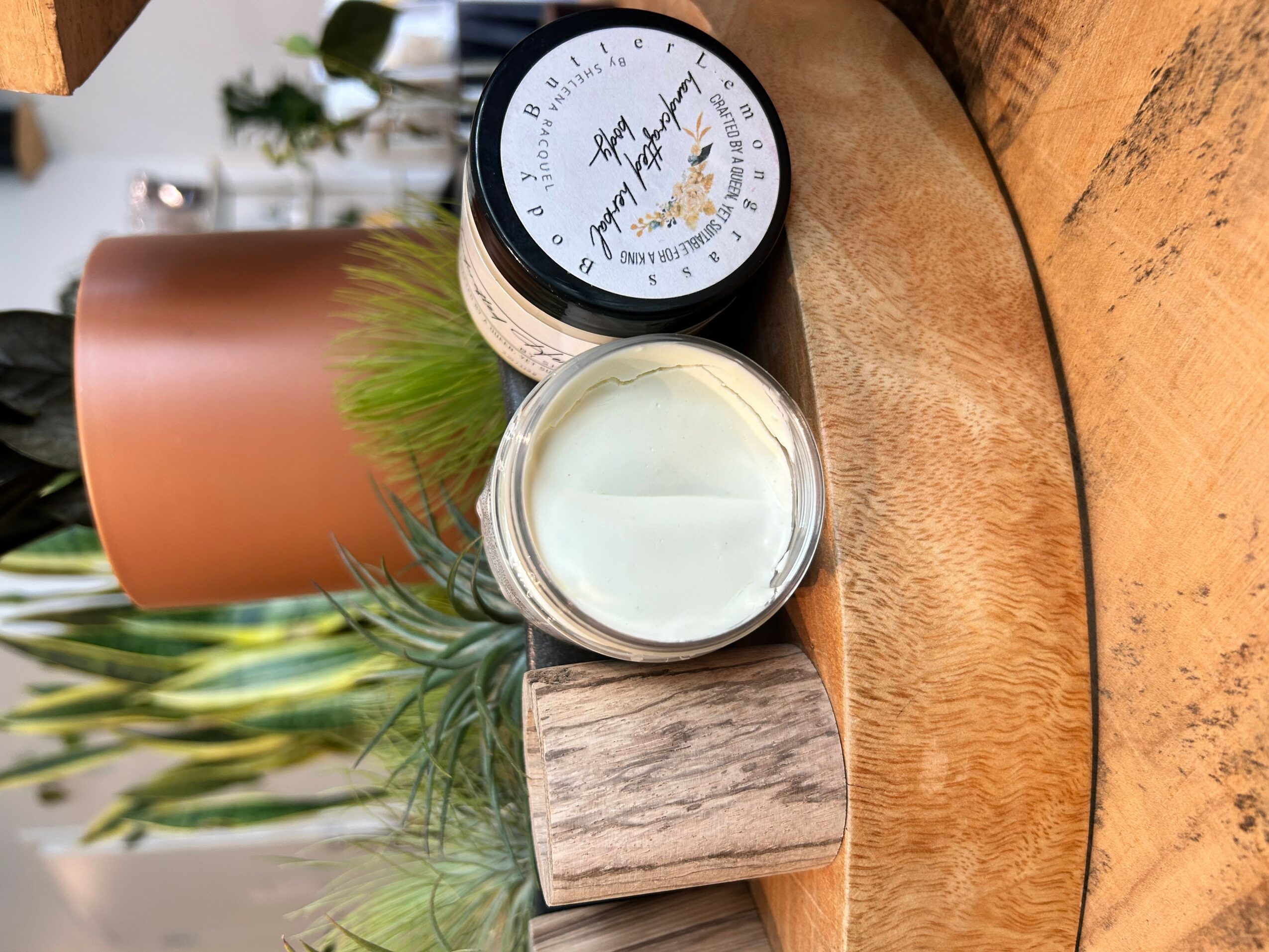 A small jar of cream, a container with a black lid, and a wooden block on a round wooden surface, with a plant in the background.