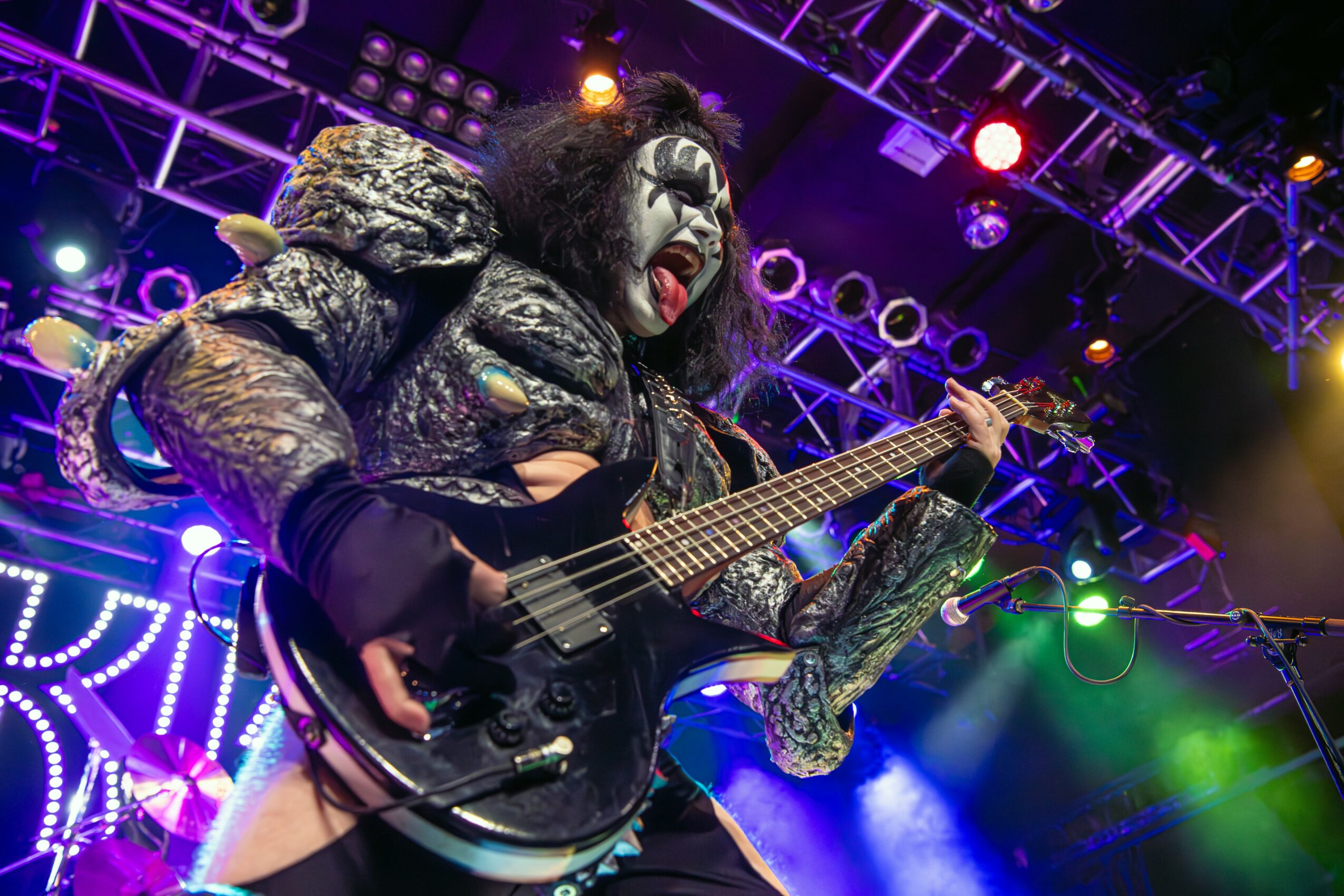 Person dressed as a rock musician with face paint, playing a guitar on stage with colorful lights.