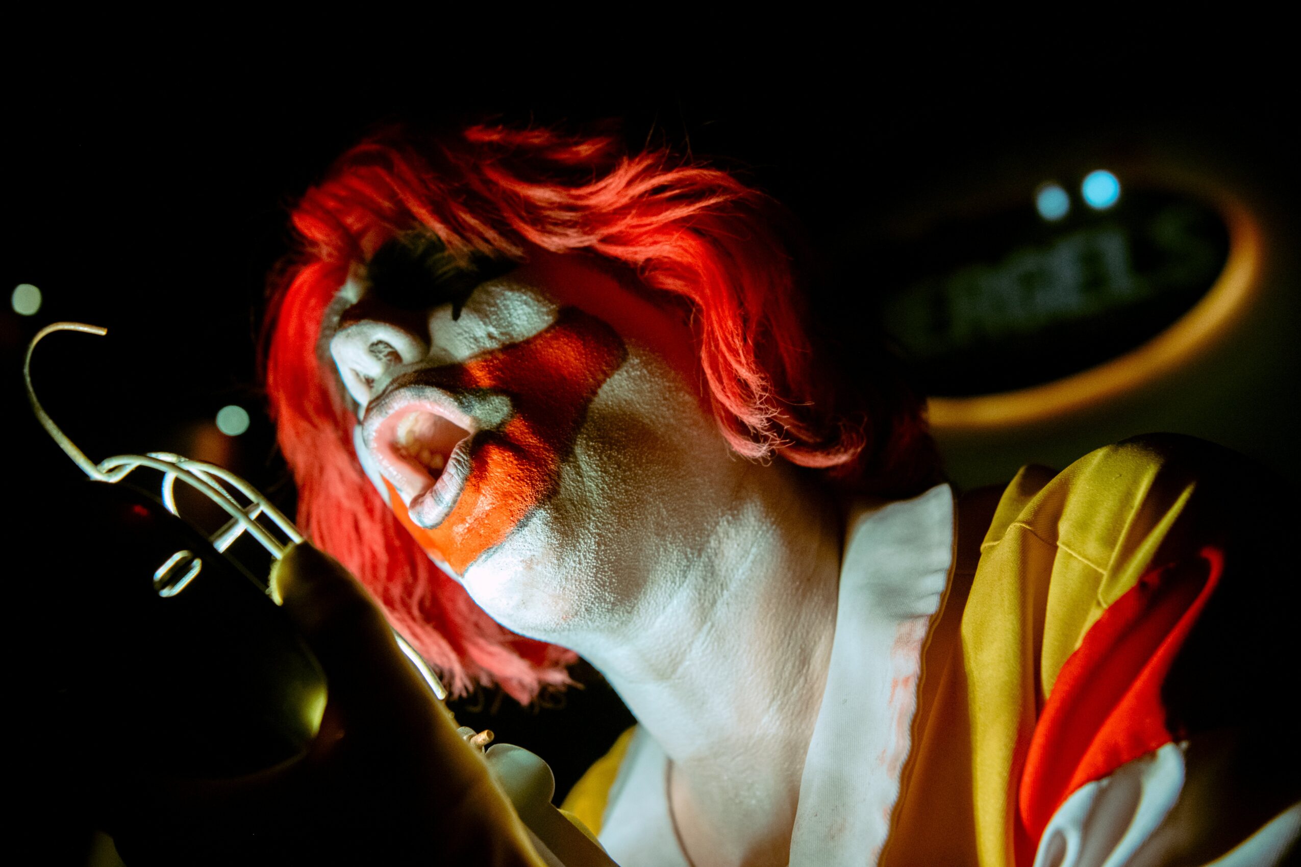 Person with red hair singing passionately, holding a microphone, wearing a yellow and red jacket, in a dark setting.