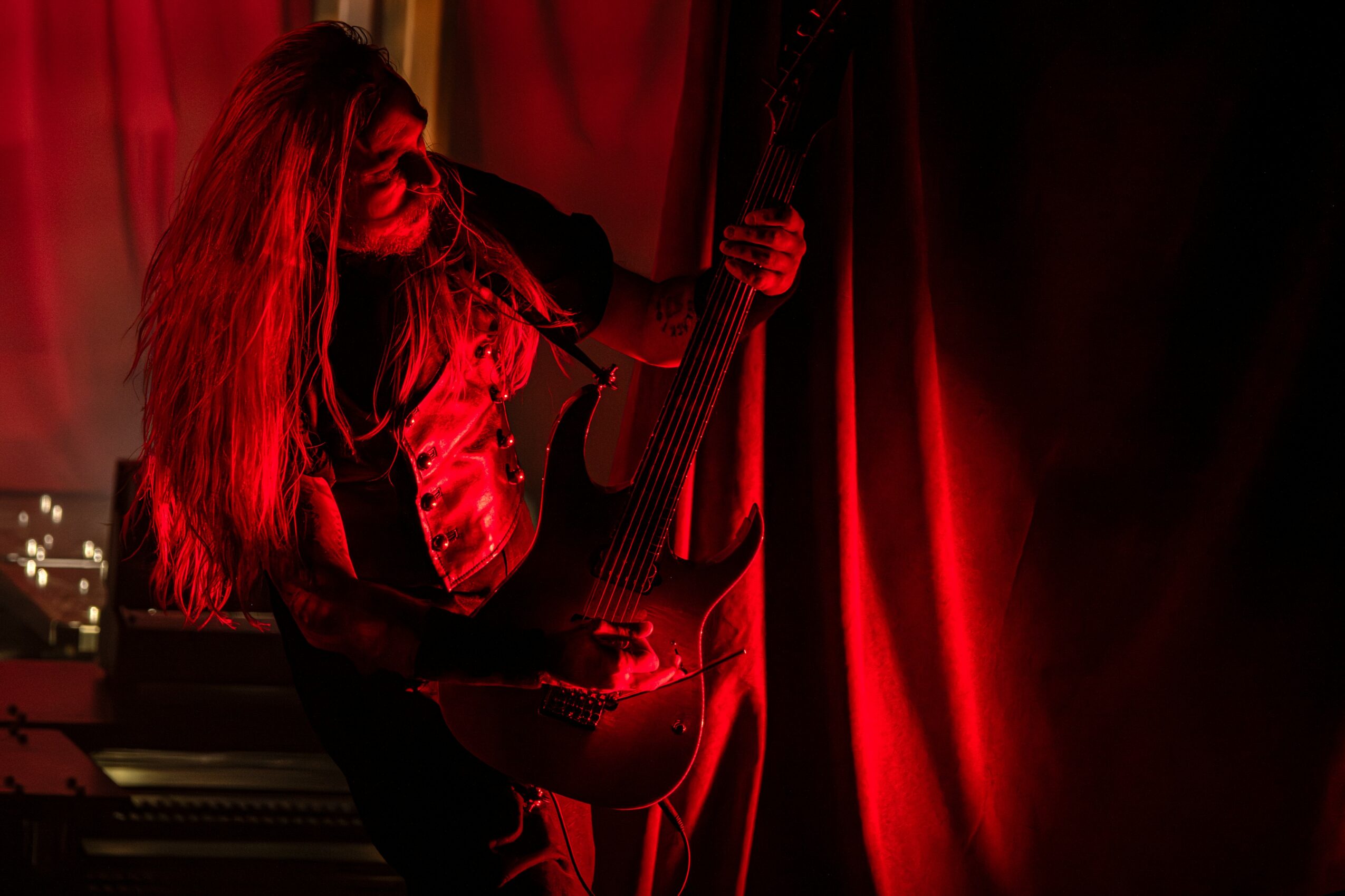 Person with long hair playing an acoustic guitar on stage with red curtains, dim lighting, and a window in the background.