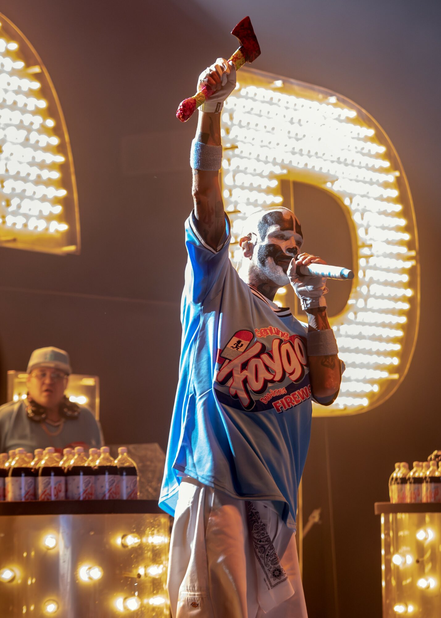 Performer on stage holding a microphone and raising a red object, with bright lights and a large illuminated letter in the background.