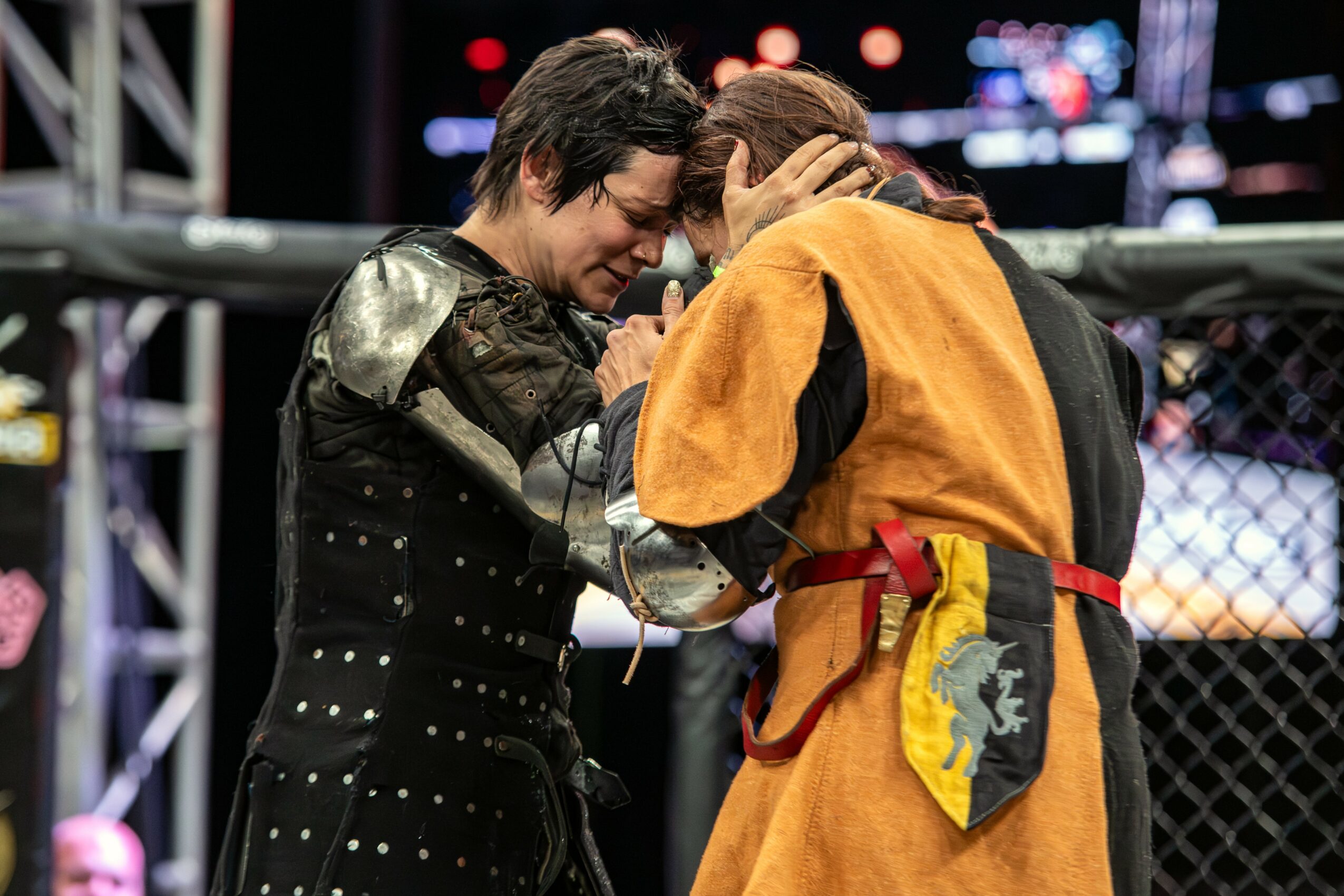 Two women in sports outfits are in a cage, touching foreheads and holding hands, with a blurred background of lights.