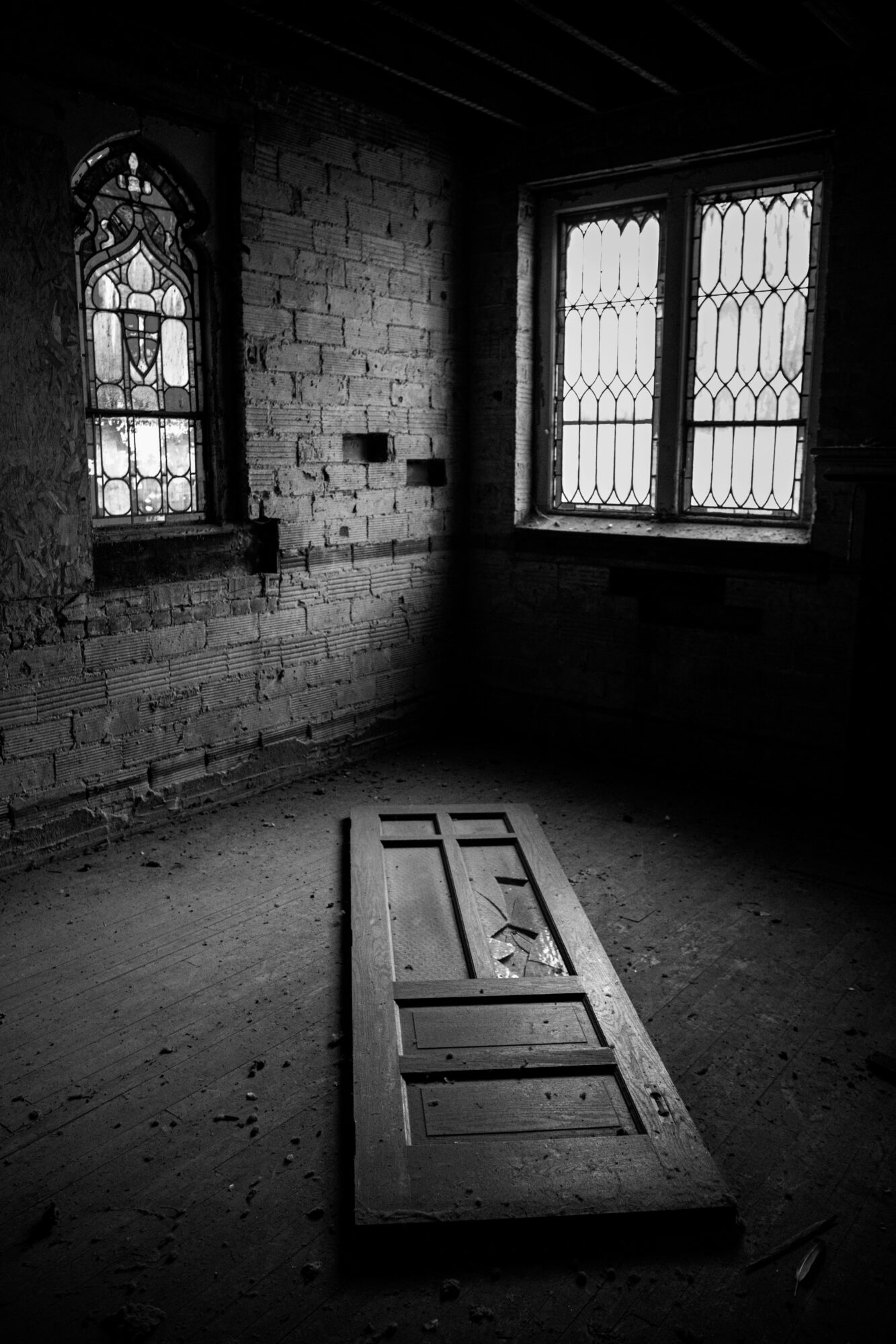 A room with brick walls, two windows with decorative glass, and a fallen wooden door on the floor.