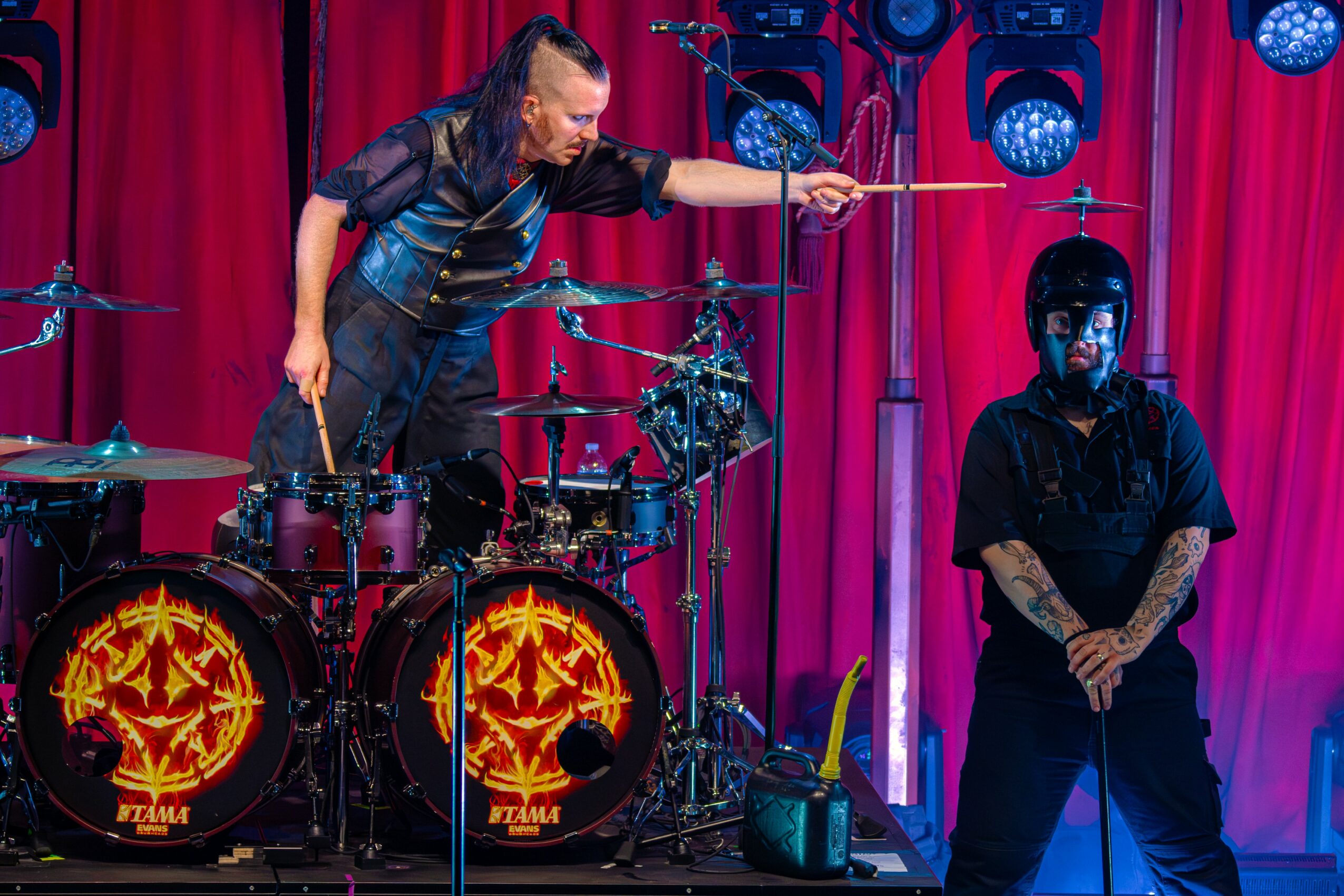Performer with drumsticks on stage, another person standing nearby, red curtain backdrop, stage lights, and musical equipment.