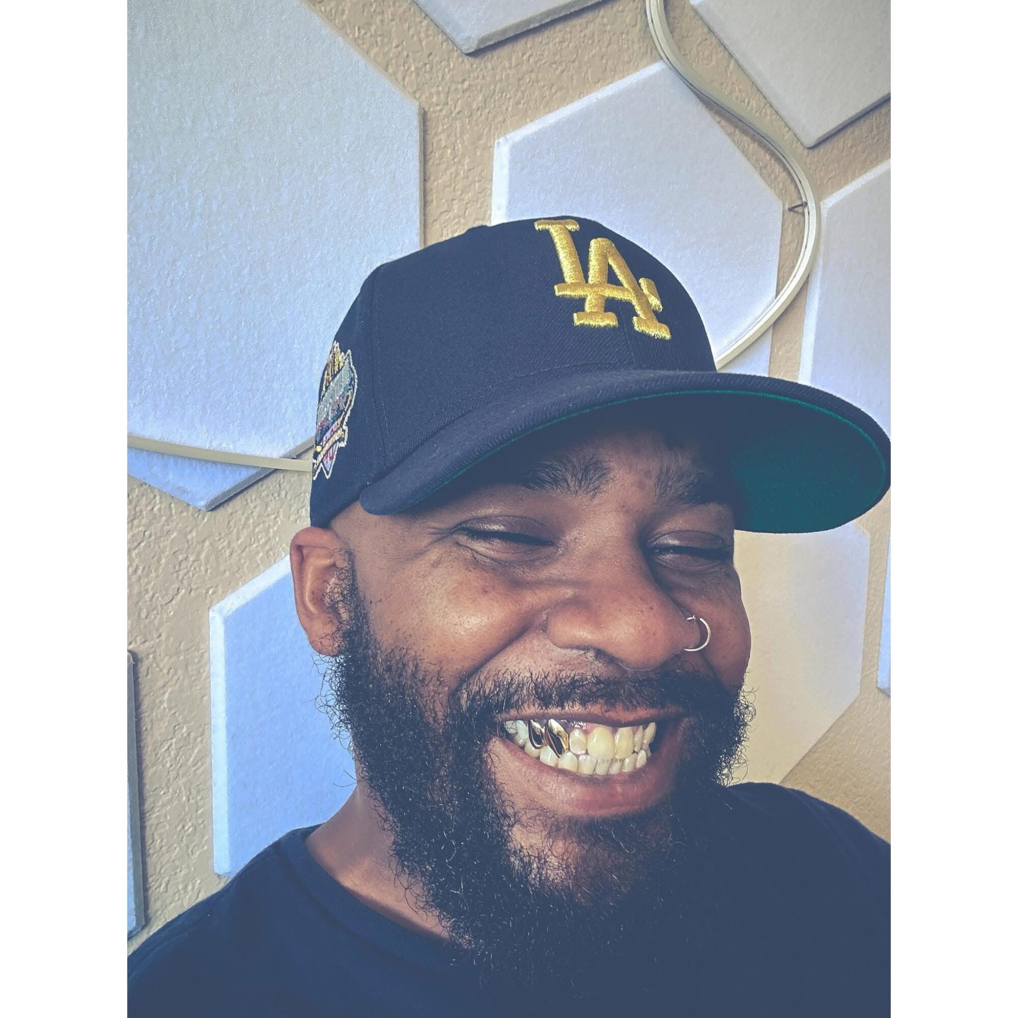 Smiling man with a beard, wearing a black LA baseball cap, showing teeth, against a textured wall background.