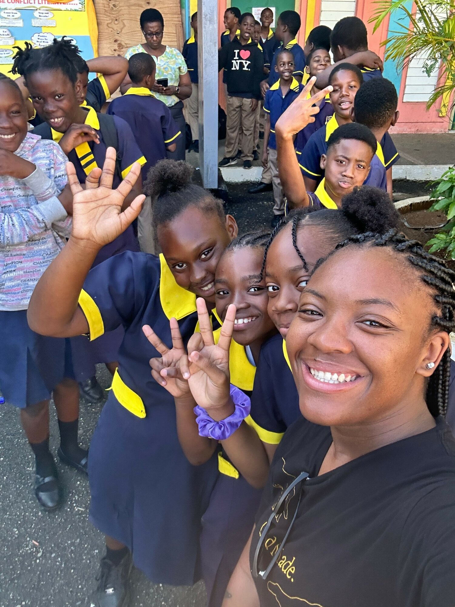 Group of children and a woman taking a selfie outdoors, children making peace signs, smiling faces, colorful background.