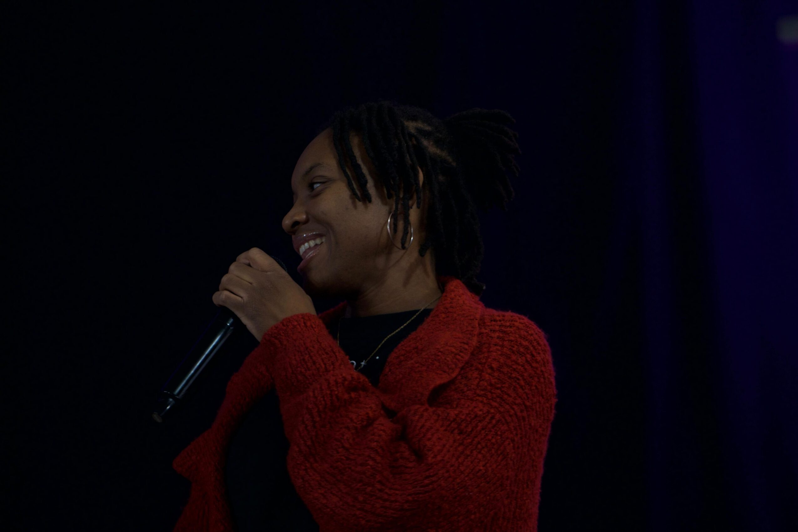 Person smiling and speaking into a microphone, wearing a red sweater, dark background.
