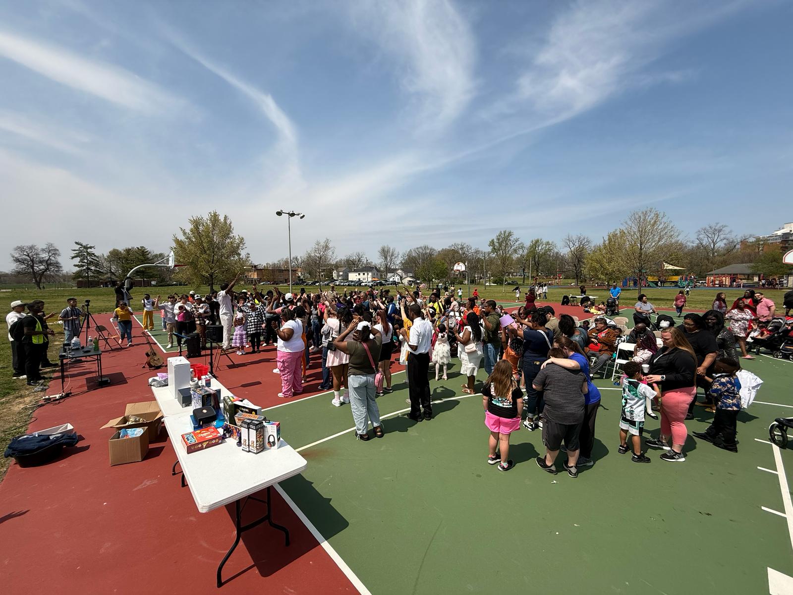 Crowd gathered on a sports court outdoors with tables and equipment, under a blue sky with clouds.