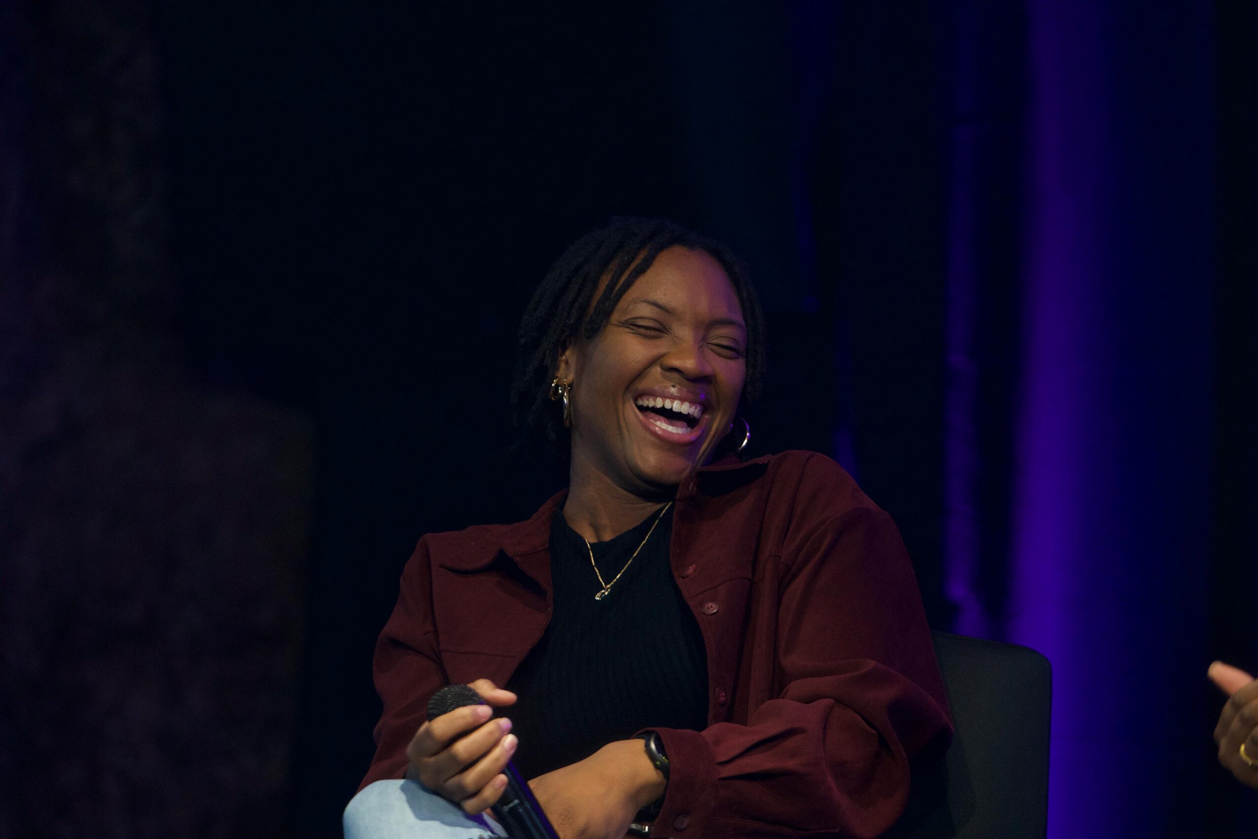 Woman laughing, wearing a dark top and maroon jacket, with dark background and purple lighting, smiling broadly.