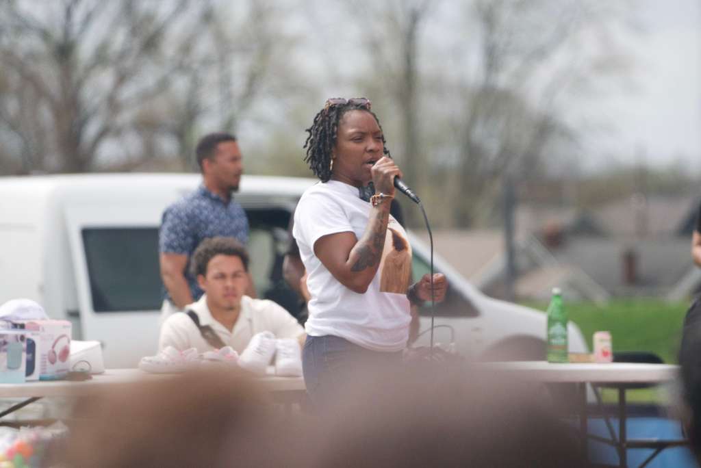 Person with dreadlocks speaking into a microphone outdoors, with people and vehicles in the background.