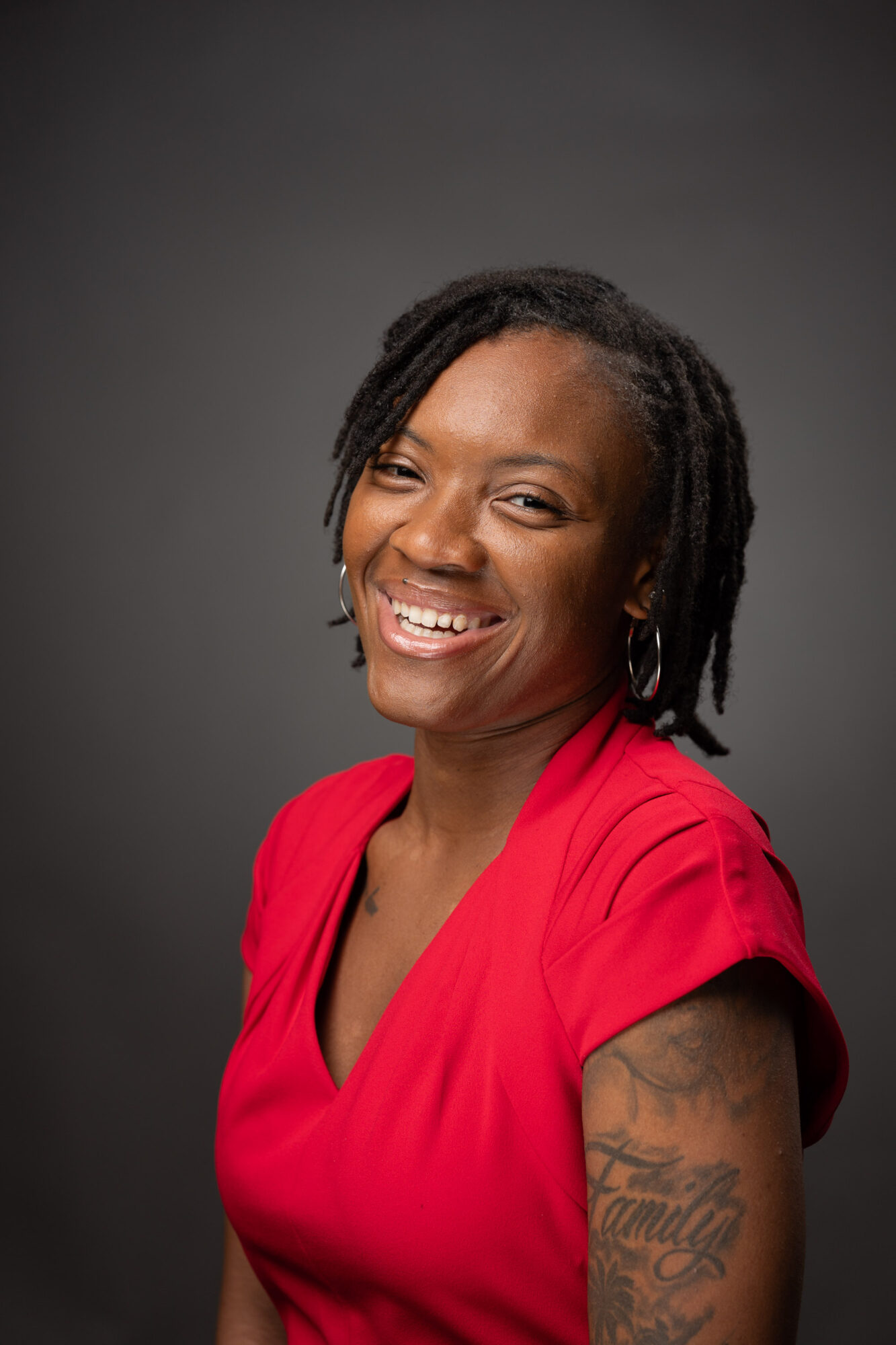 Smiling woman with short dreadlocks wearing a red top, showing a tattoo on her arm, against a dark background.