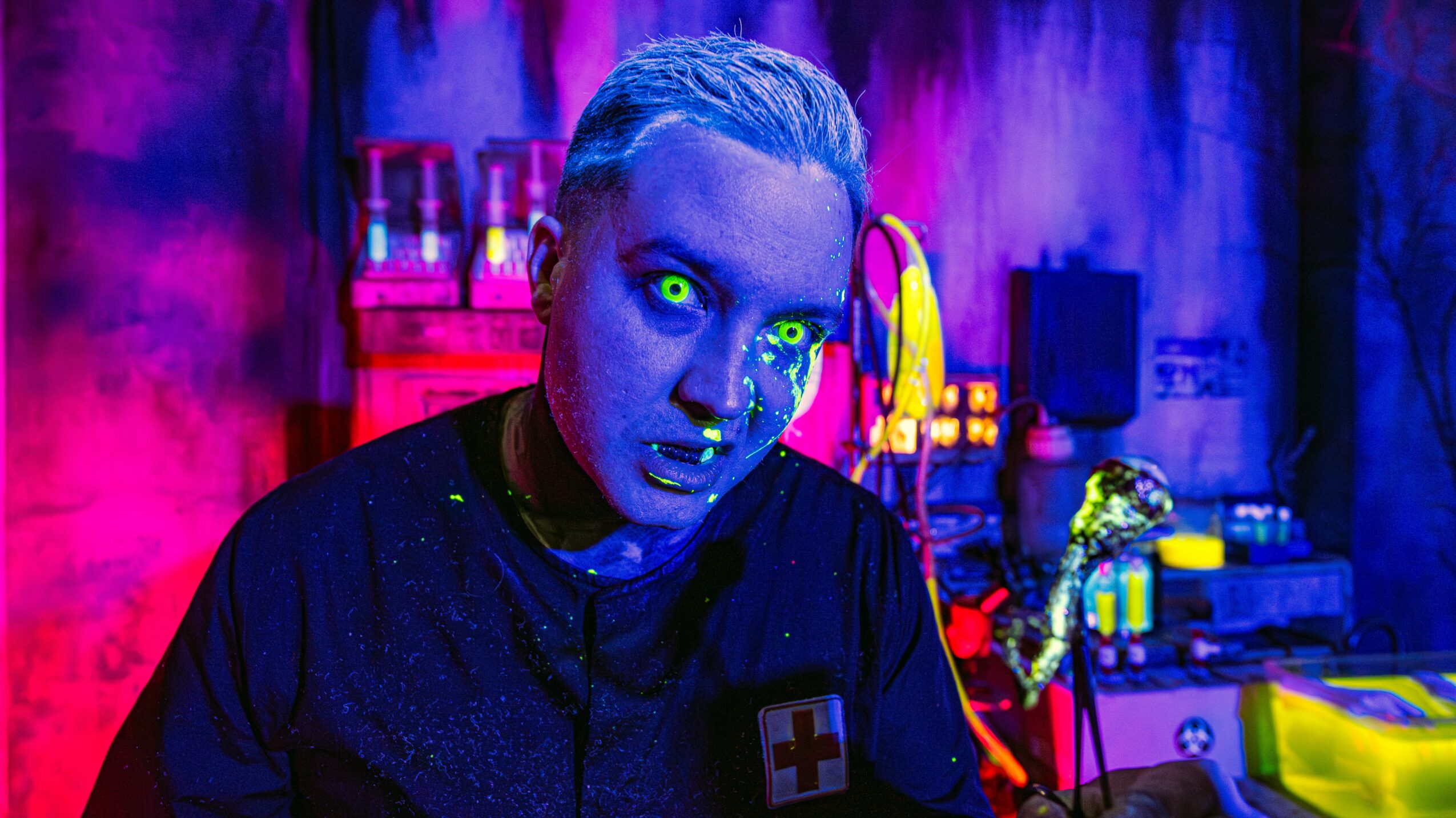 Person with blue skin and glowing green eyes in a colorful, neon-lit room, wearing a dark shirt with a red cross badge.
