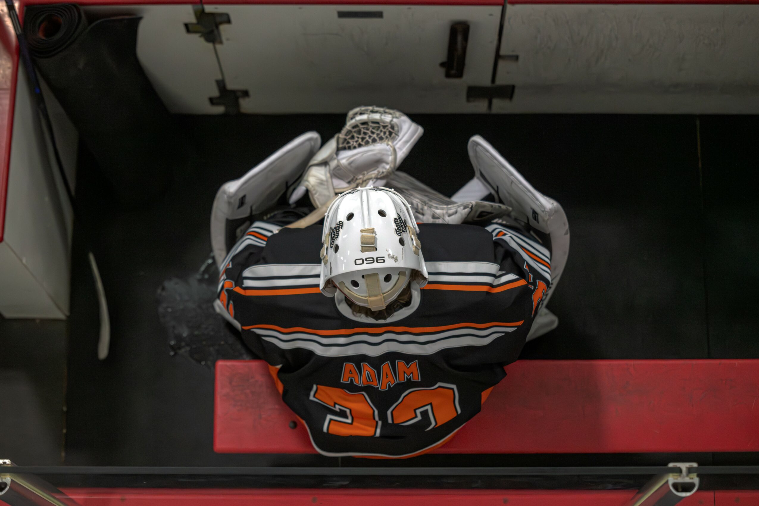 Top-down view of a hockey goalie in a black jersey with orange and white stripes, number 30, and the name 'ADAM'.
