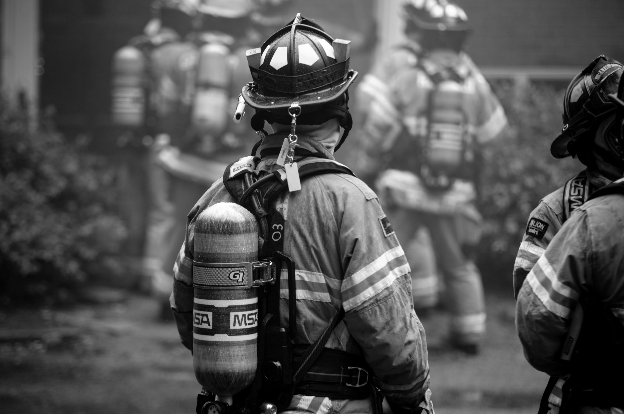 Firefighter in gear with helmet and oxygen tank, facing away, with other firefighters in background