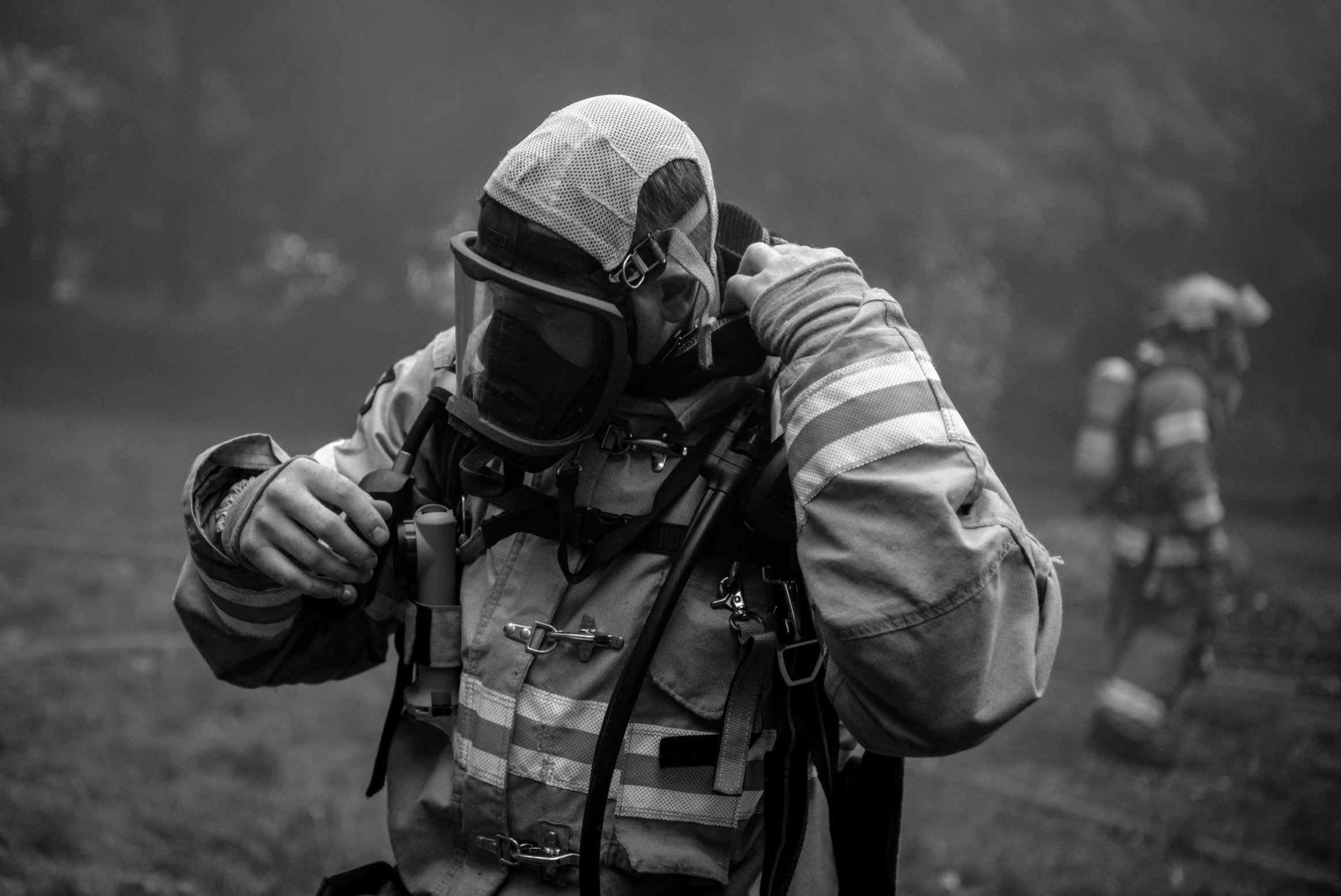 Firefighter in protective gear adjusting helmet, with other firefighters in background outdoors, black and white image.