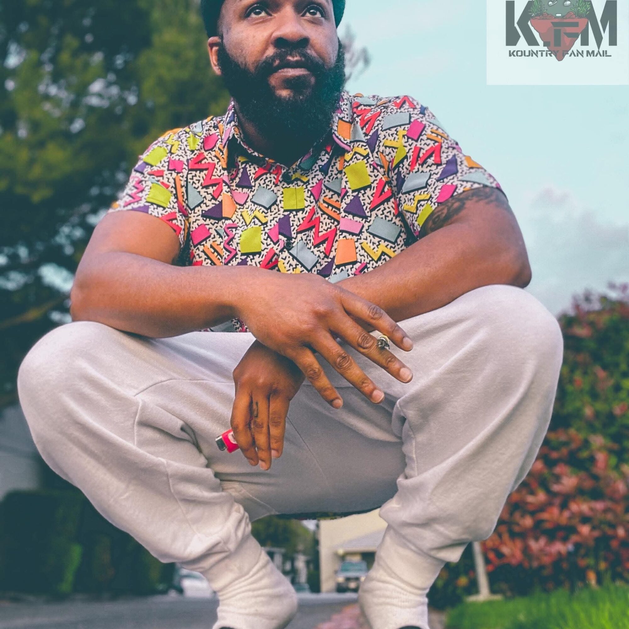 Person crouching outdoors, wearing a colorful patterned shirt and light-colored pants, with trees and sky in background.