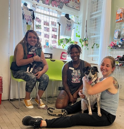Three women sitting indoors with a dog, smiling, in a room with colorful decorations and large windows.
